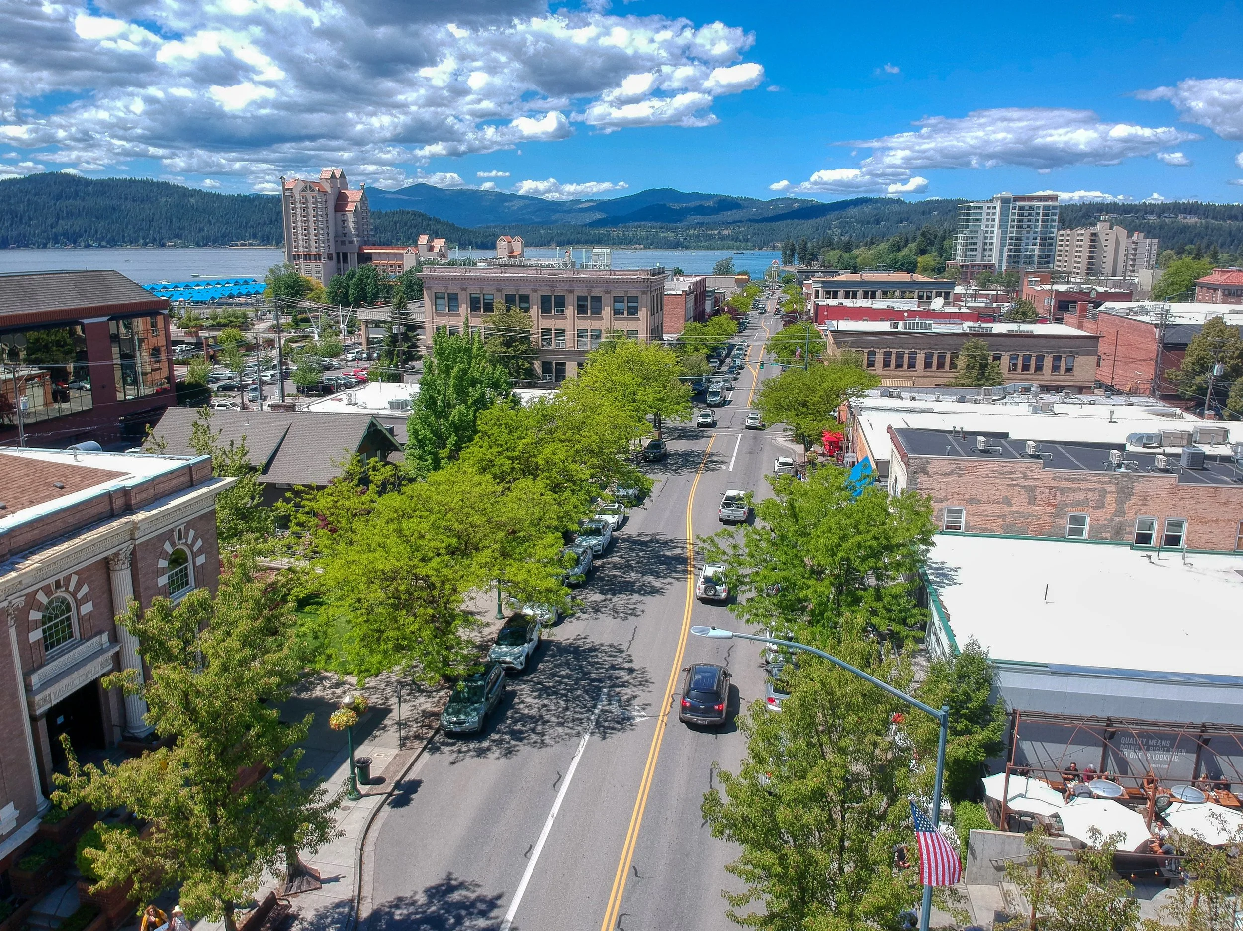 A scenic view of a small town with a street lined with trees and parked cars, buildings including brick and modern structures, and a lake with islands and a blue sky with clouds in the background.
