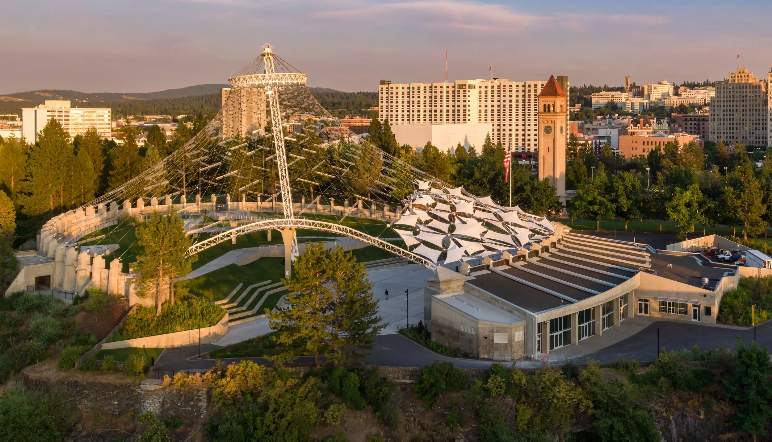 Aerial view of a city park with a modern amphitheater featuring a large white tent-like canopy and a tall, metal sculpture. Surrounding buildings include a historic clock tower, office buildings, and residential structures. Trees and greenery are visible throughout the park and cityscape, with a hills in the background and a sunset sky.