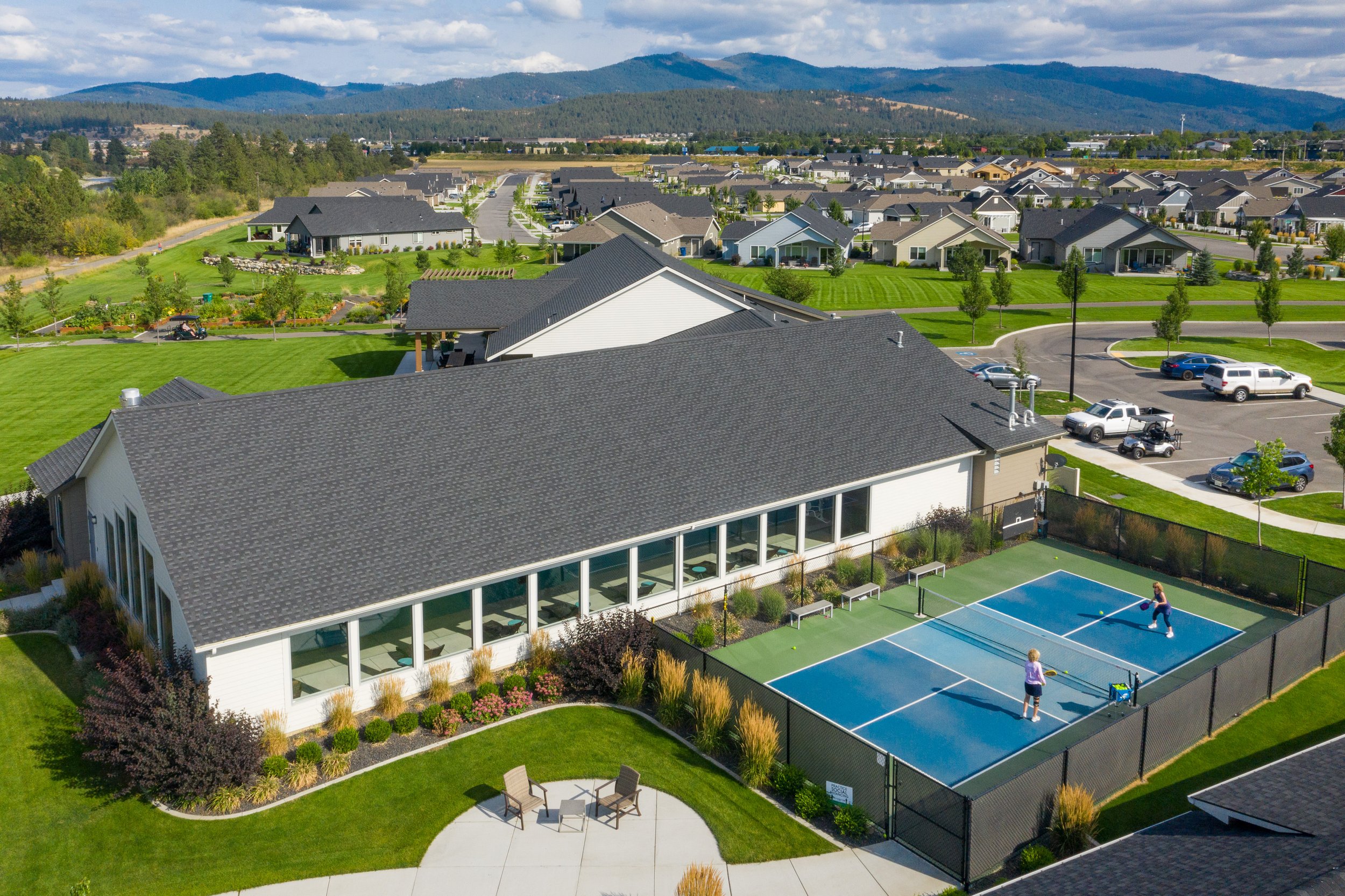 Aerial view of a residential neighborhood with houses, green lawns, a tennis court with two people playing, parking lots with cars, and distant mountains under partly cloudy skies.
