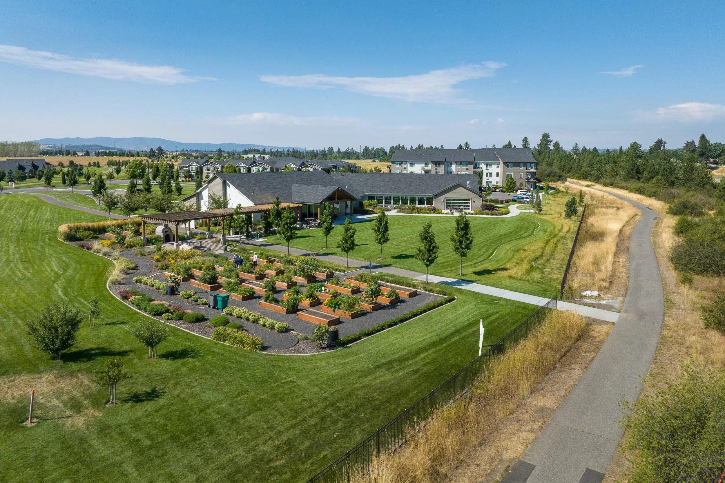 A landscaped park with green grass, young trees, and a garden area infront of residential buildings under a blue sky.