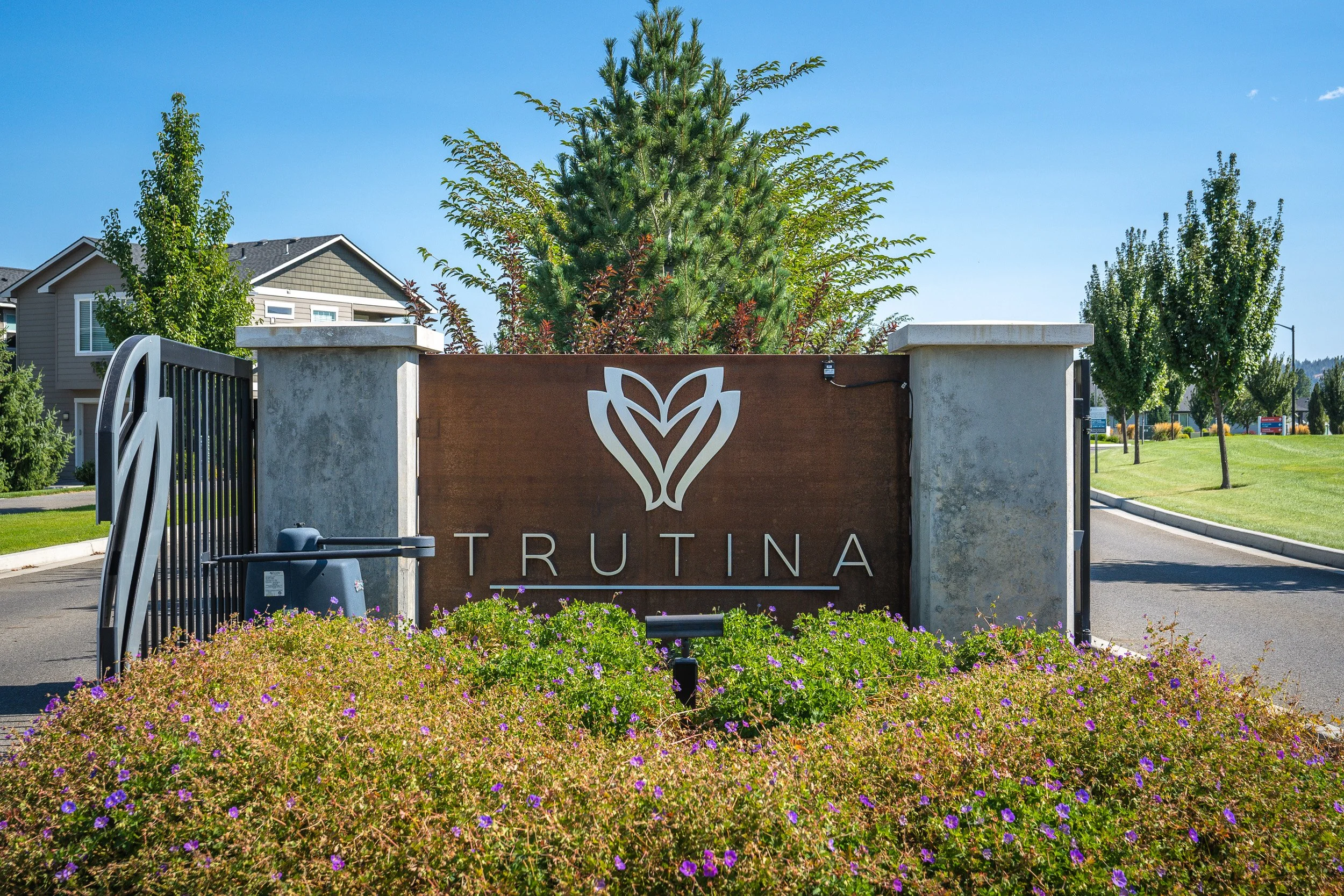 Entrance sign for Trutina with a logo and a landscaped area with purple flowers in the foreground and trees in the background.