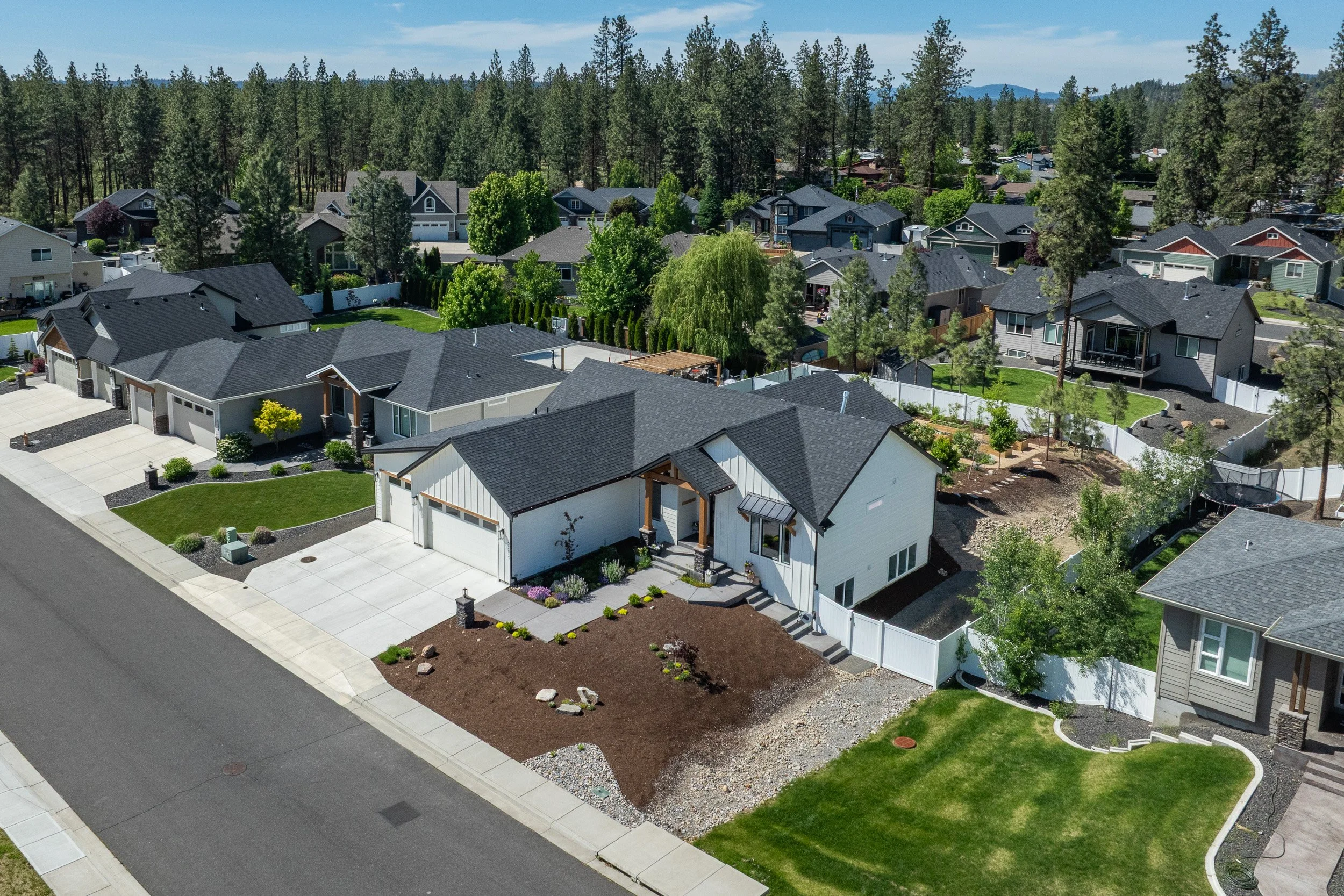 Aerial view of a suburban neighborhood with modern houses, green lawns, trees, and a forest in the background.