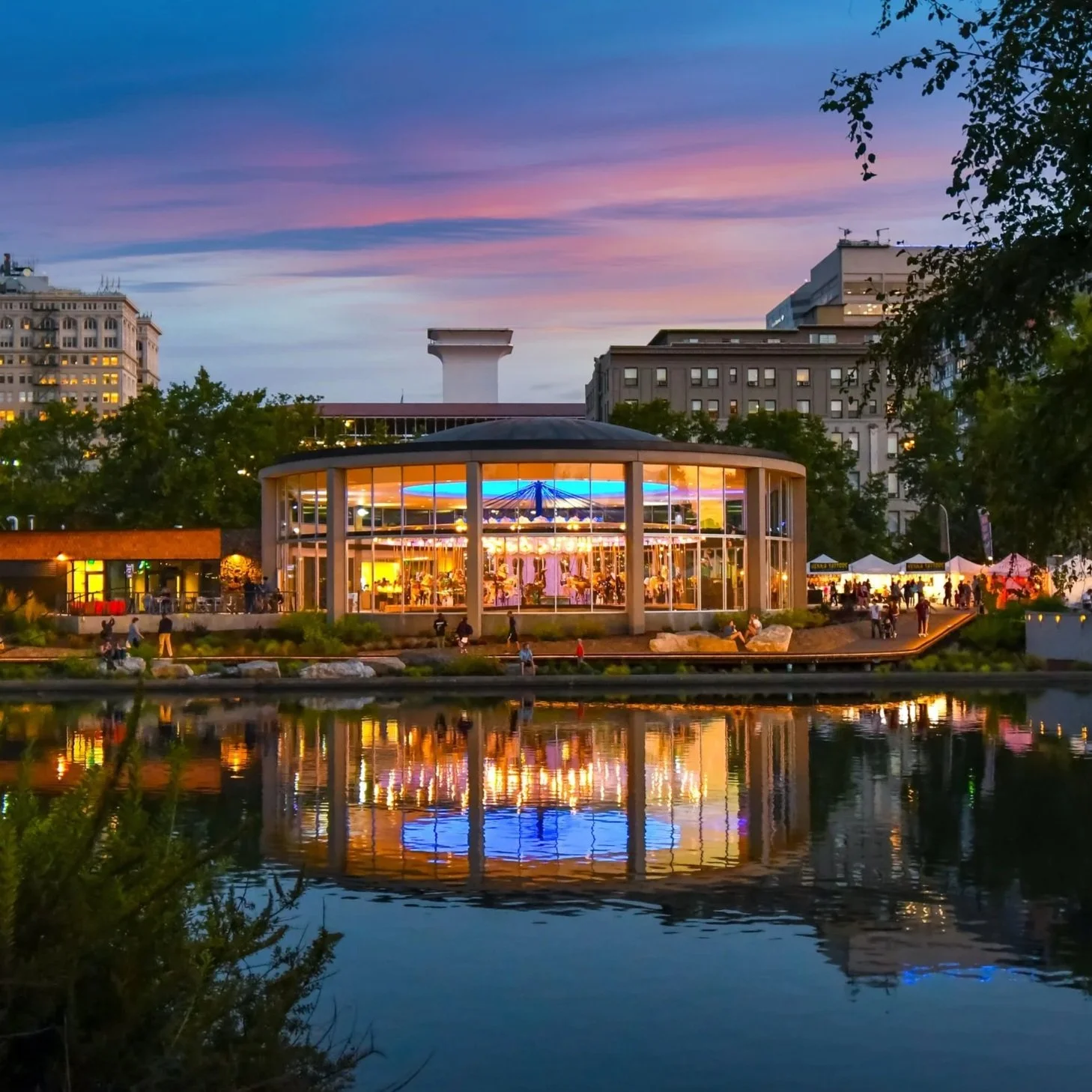 A waterfront view of a city park during sunset, featuring a lit-up glass pavilion with people inside and outside, with trees, buildings, and a colorful sky reflected in the water.