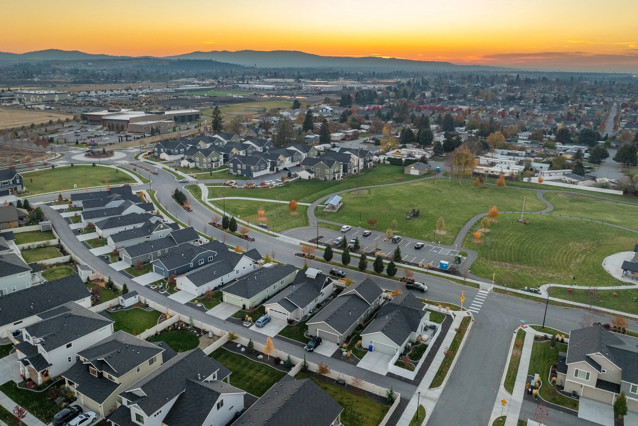 An aerial view of a suburban neighborhood during sunset, showing houses with yards, streets, a park with walking paths, and distant industrial areas and mountains.