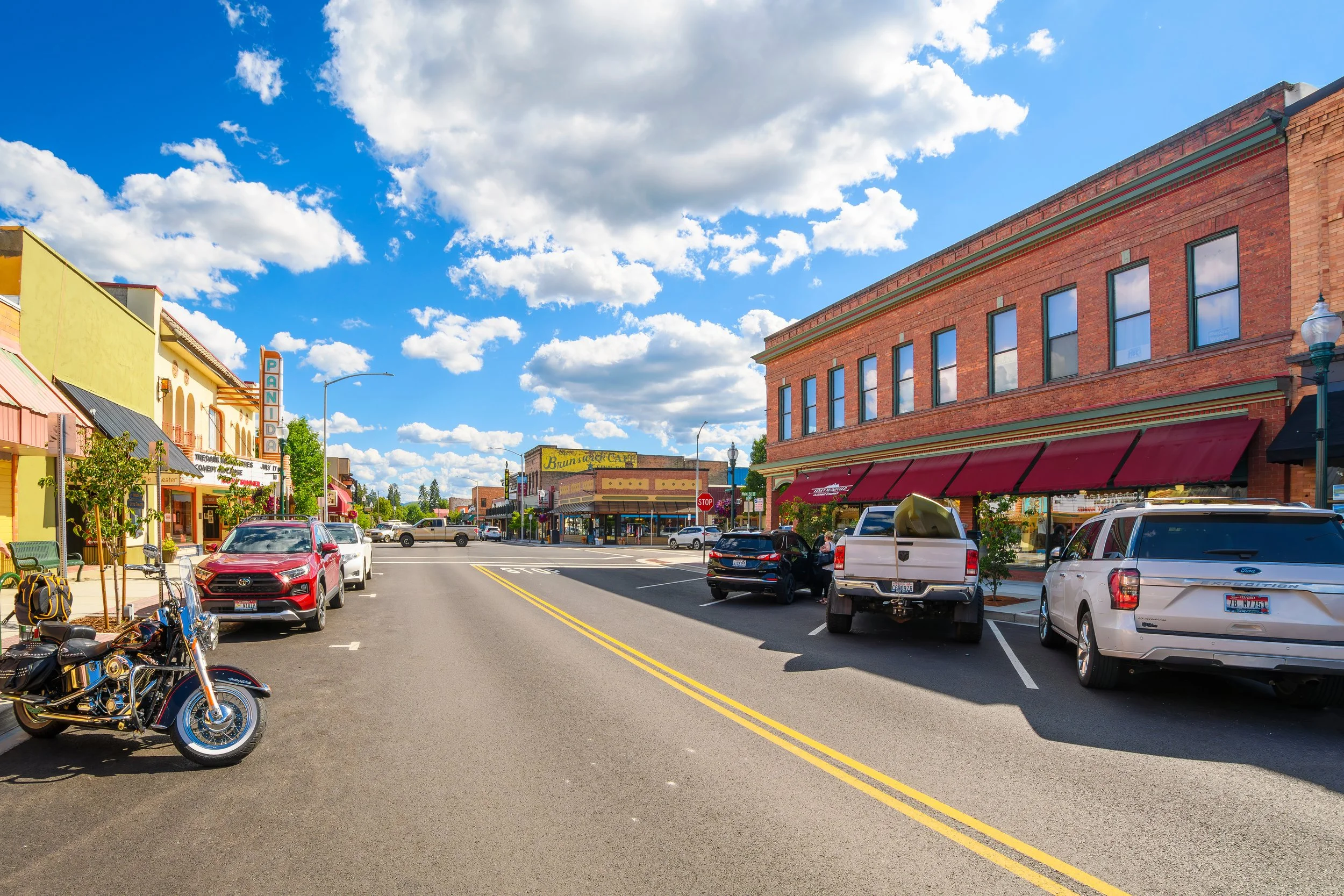 Street scene with cars and motorcycles parked along the sidewalk, brick buildings with awnings, trees, street signs, and a blue sky with scattered clouds.