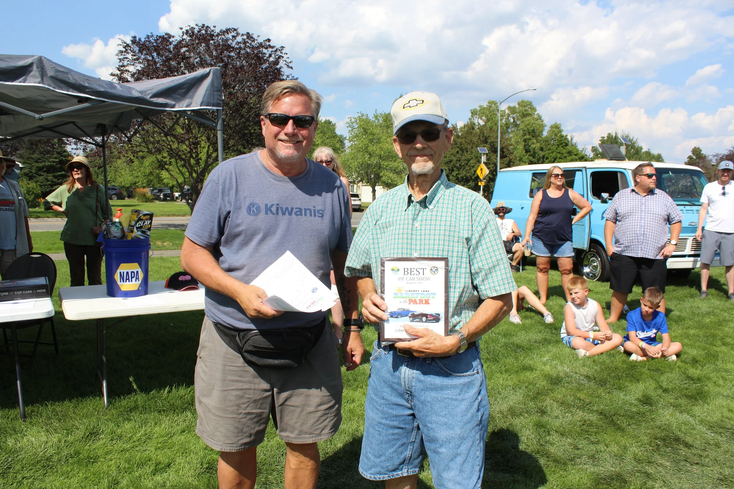 Real Estate Agent - Tim Olsen outdoors at a car show, one holding an award and the other holding documents. Several people and vintage vehicles are visible in the background under a partly cloudy sky.
