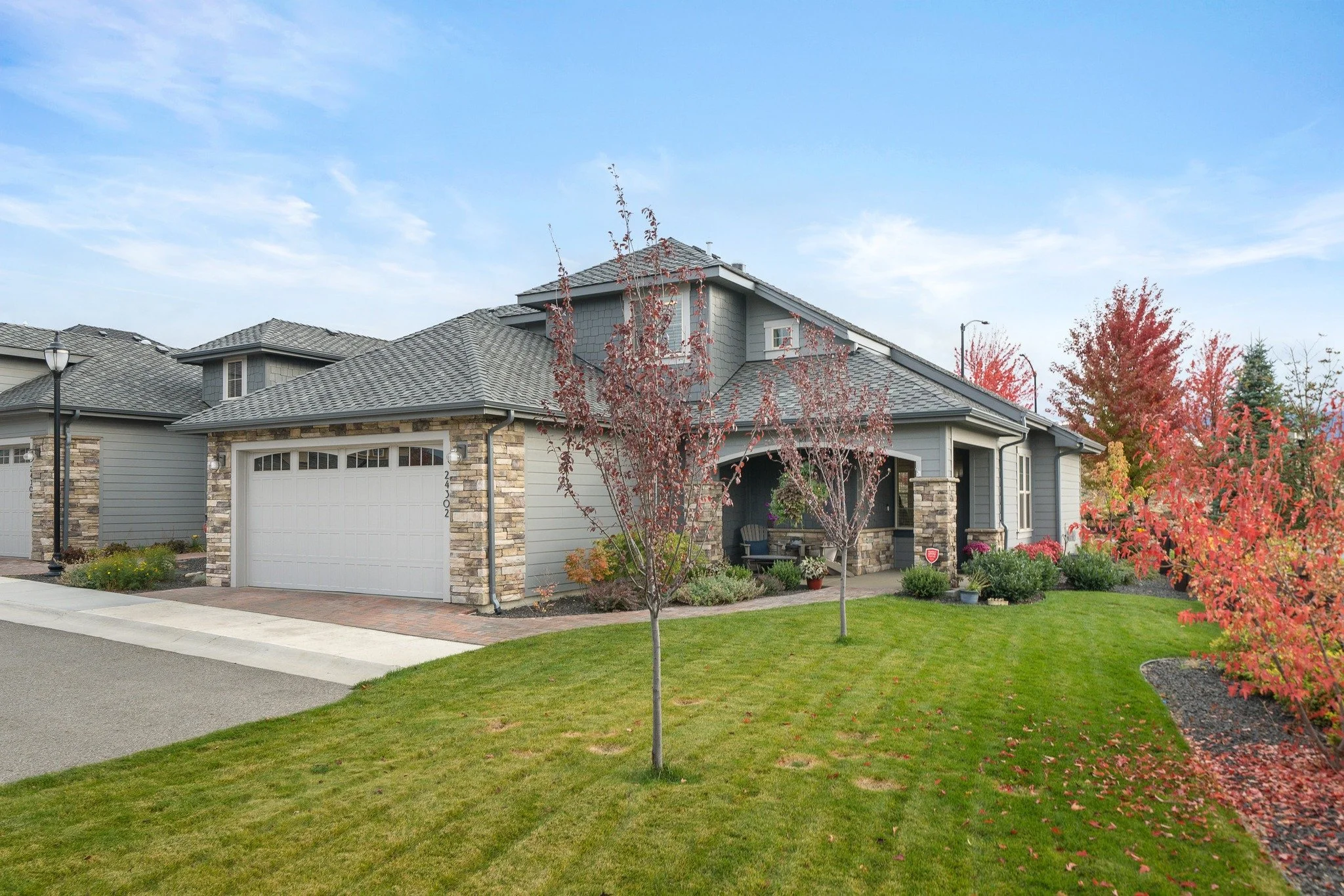 A suburban house with a gray roof, stone and siding exterior, a white garage door, and a front porch with two chairs. The yard has a young tree with red leaves, green grass, and flower beds with autumn foliage. The sky is mostly clear with some wispy clouds.