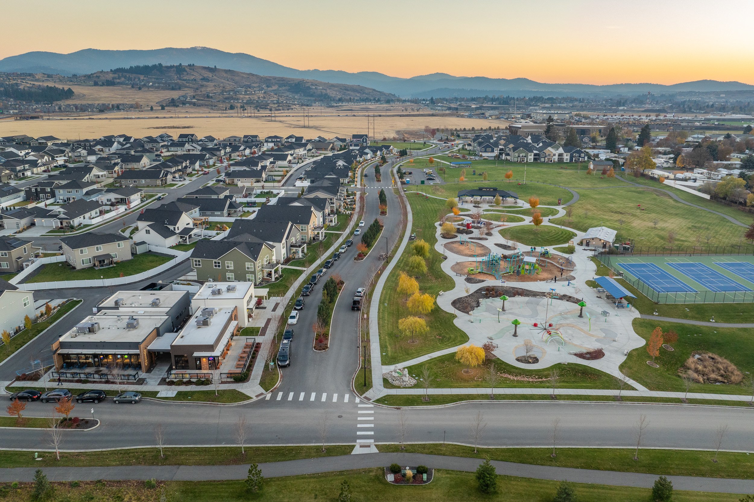 Aerial view of a suburban neighborhood with houses, a park with playground equipment, tennis courts, and parking lots, set against rolling hills and mountains at sunset.