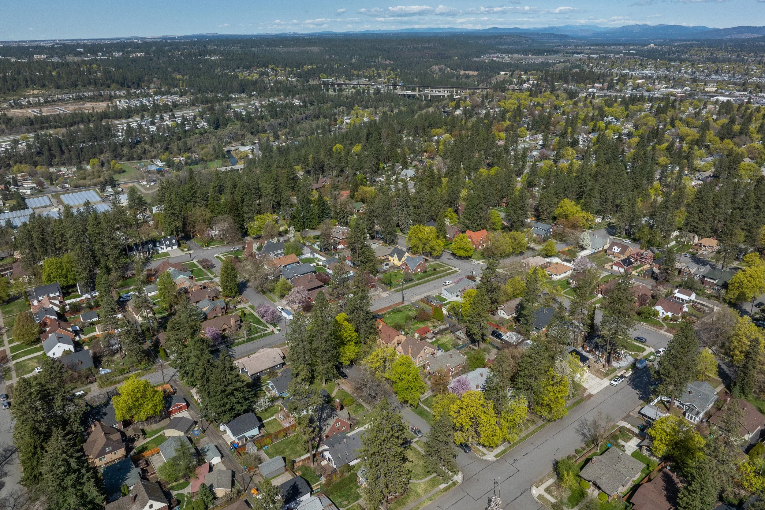 An aerial view of a suburban neighborhood with houses, trees, and streets, with a forested area and mountains in the distance under a partly cloudy sky.
