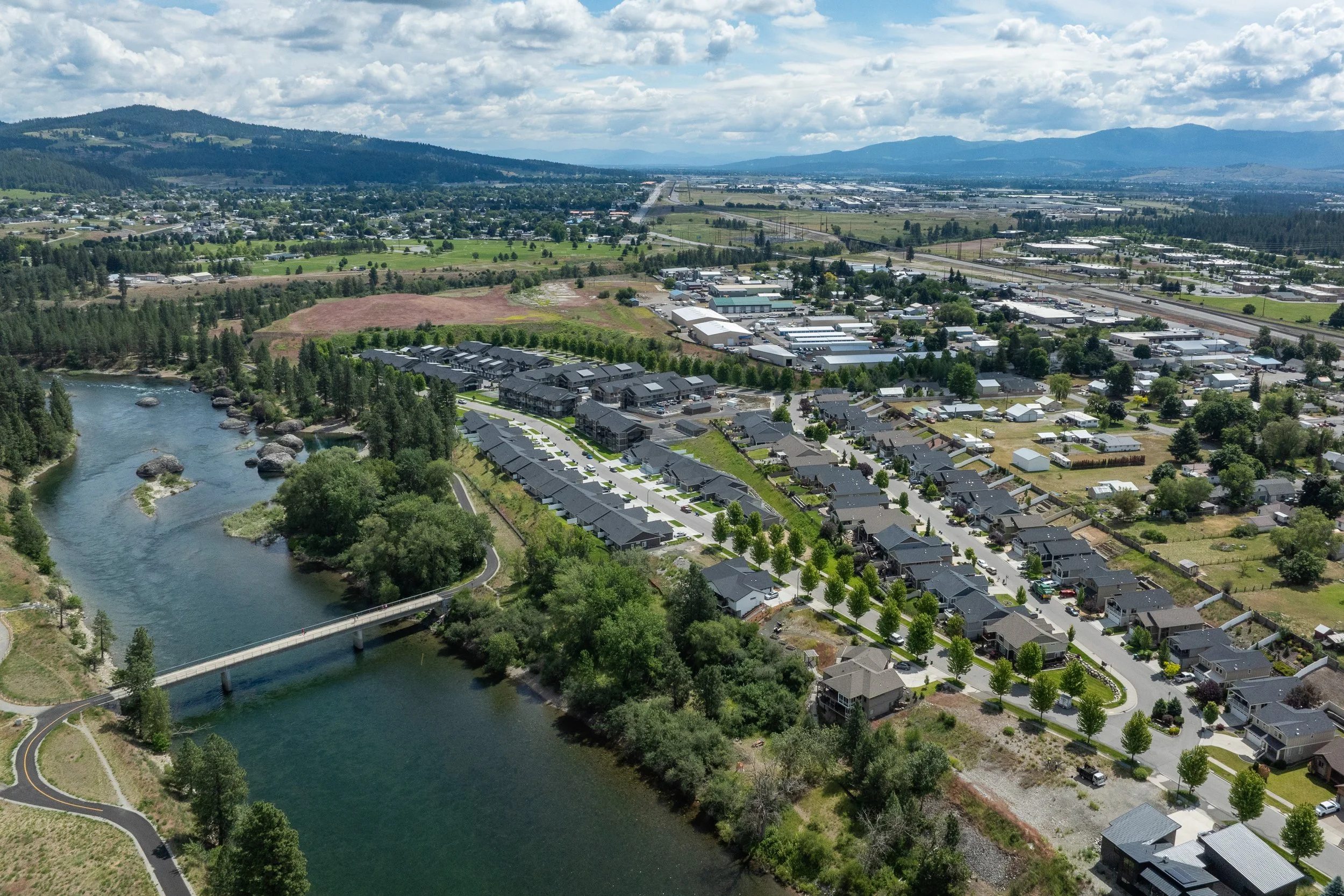 Aerial view of a river flowing through a residential and commercial area with houses, trees, roads, and mountains in the background under a partly cloudy sky.