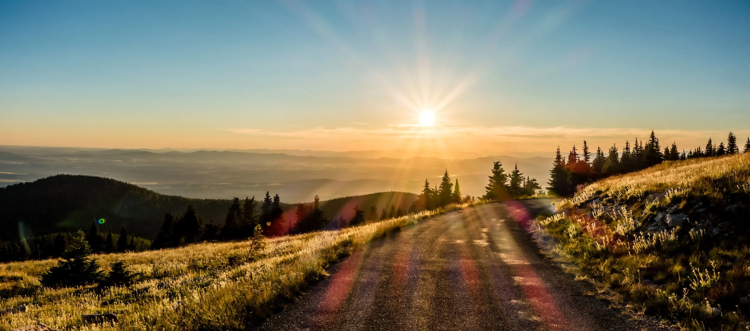 A scenic landscape of a mountain road at sunrise, with the sun illuminating a forested hillside and distant misty mountains.