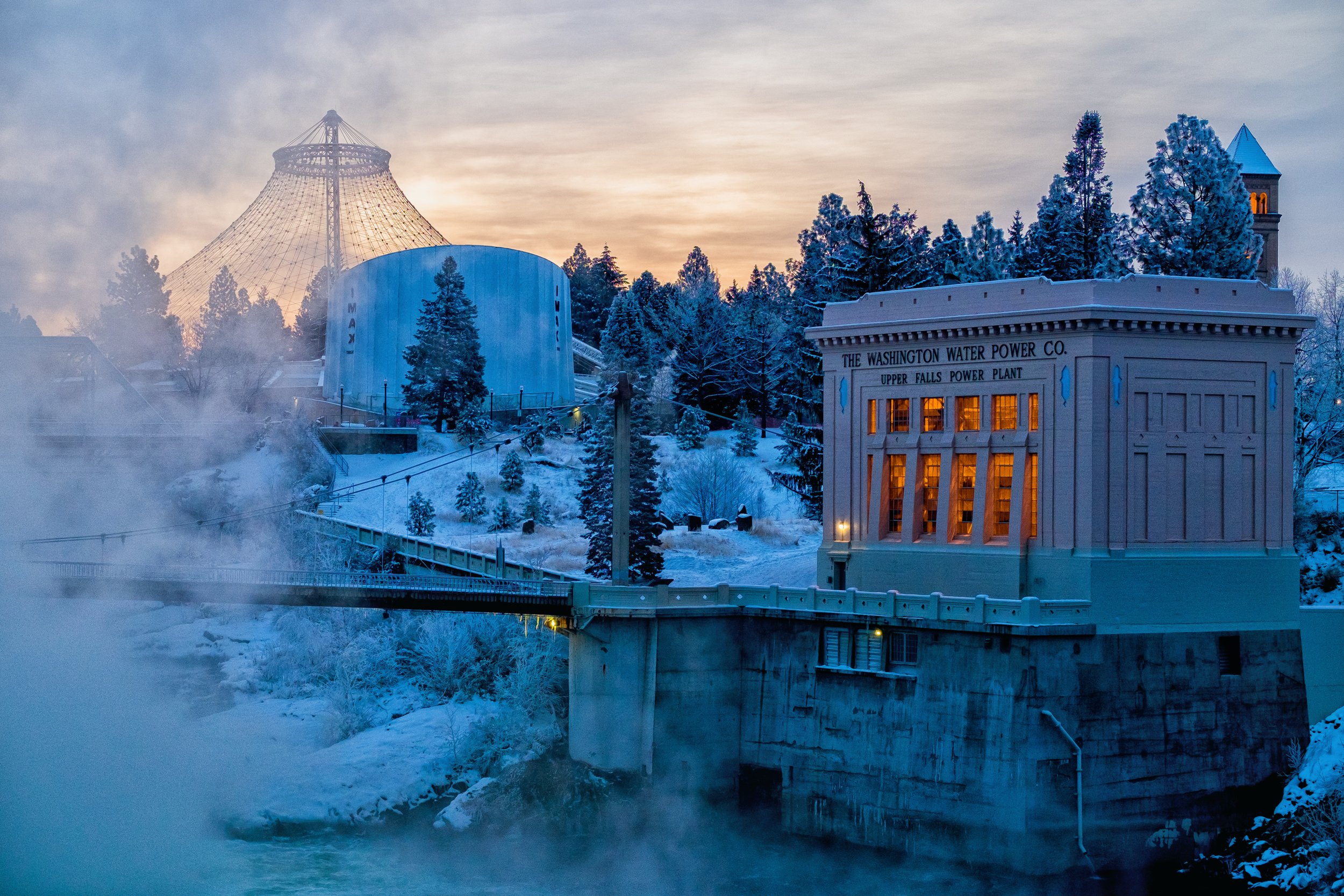 View of a snow-covered dam and hydroelectric power plant with steam rising from the water, surrounded by snow-blanketed trees during dusk.