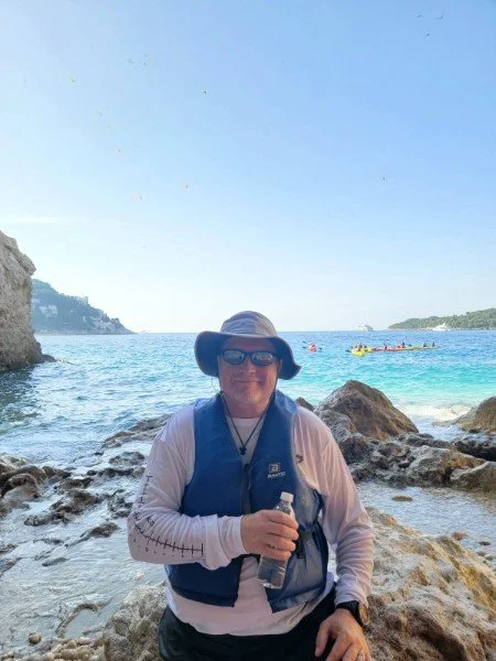 Real Estate Agent - Tim Olsen wearing a hat and sunglasses, standing on rocks at the seaside with boats and distant land in the background.