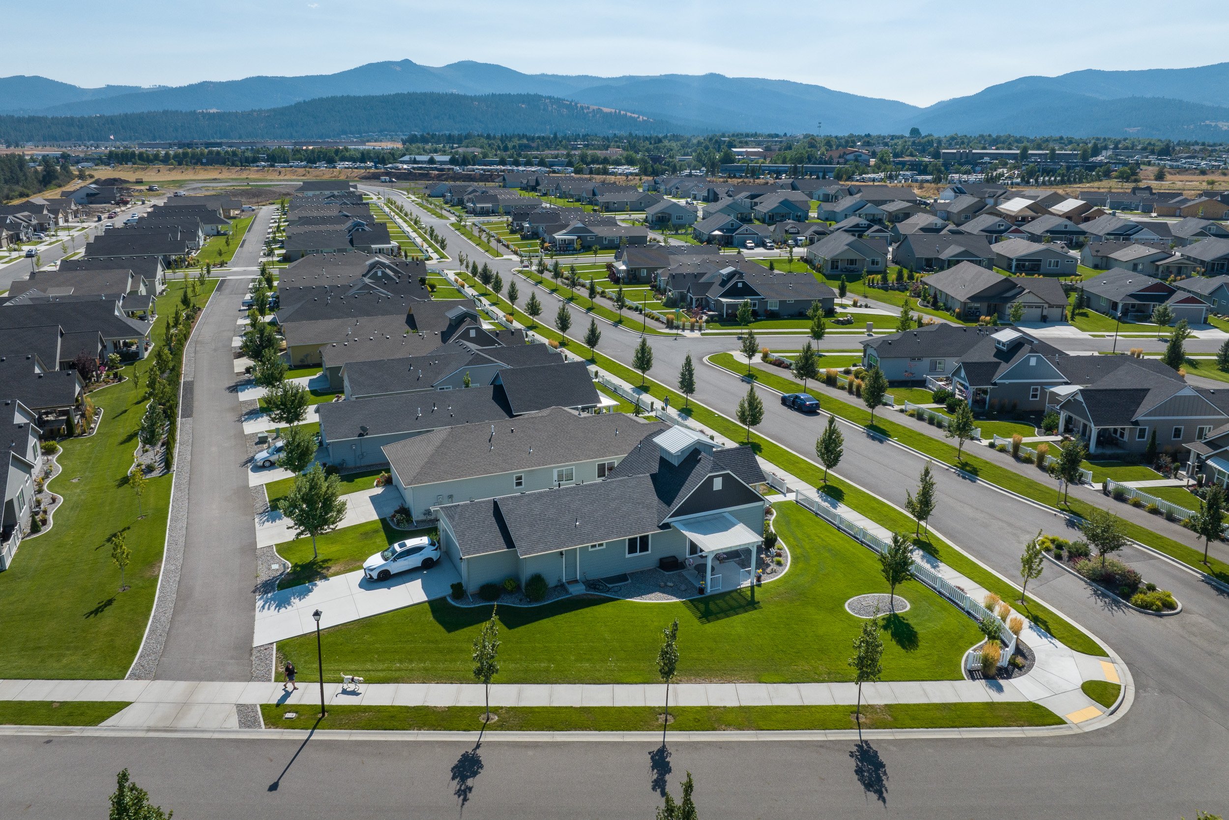 An aerial view of a suburban neighborhood with rows of houses, green lawns, and tree-lined streets, with mountains in the background under a cloudy sky.
