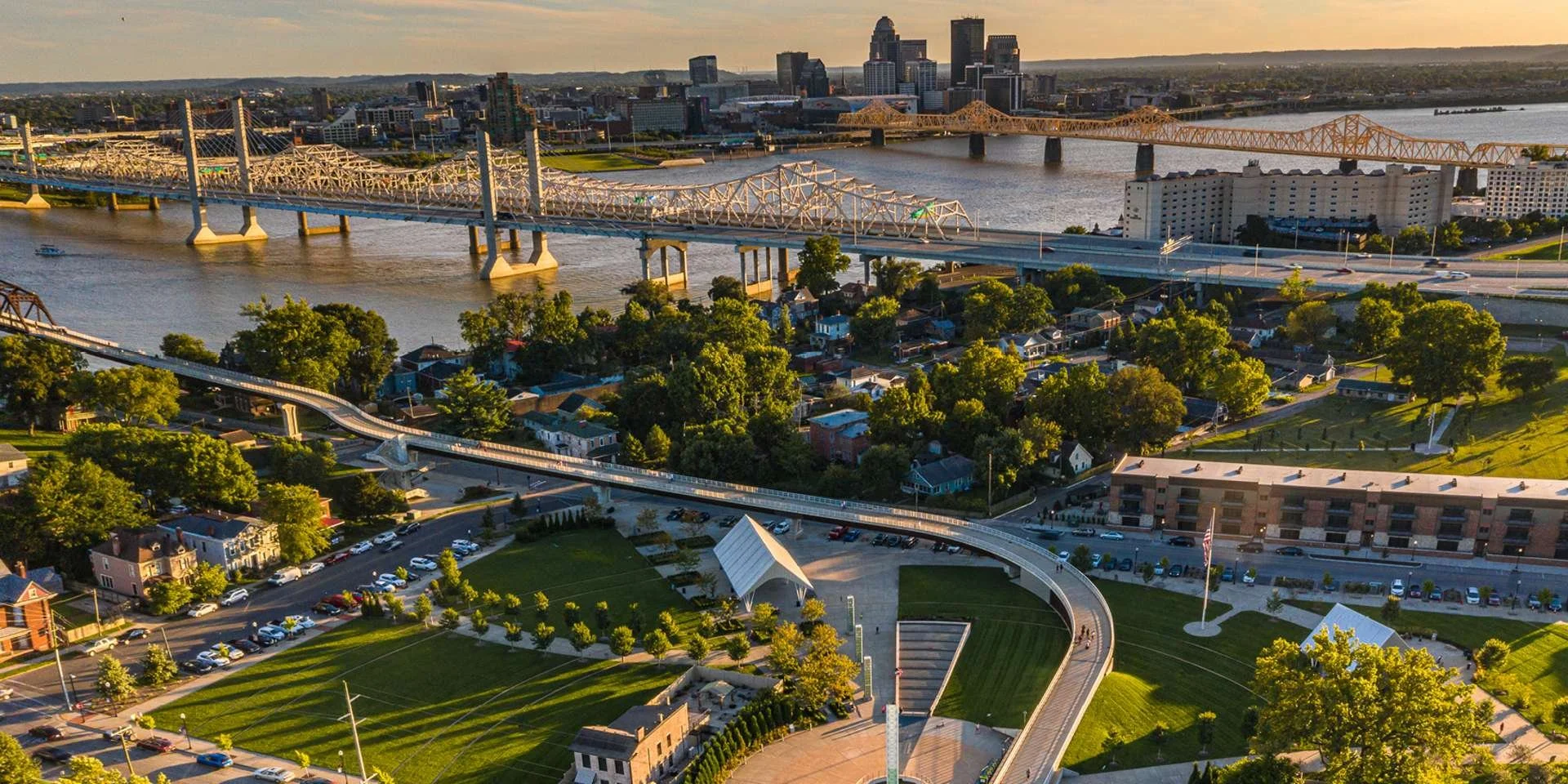 A cityscape with bridges over a river, residential houses, a park with a flag, and downtown buildings in the background during sunset.