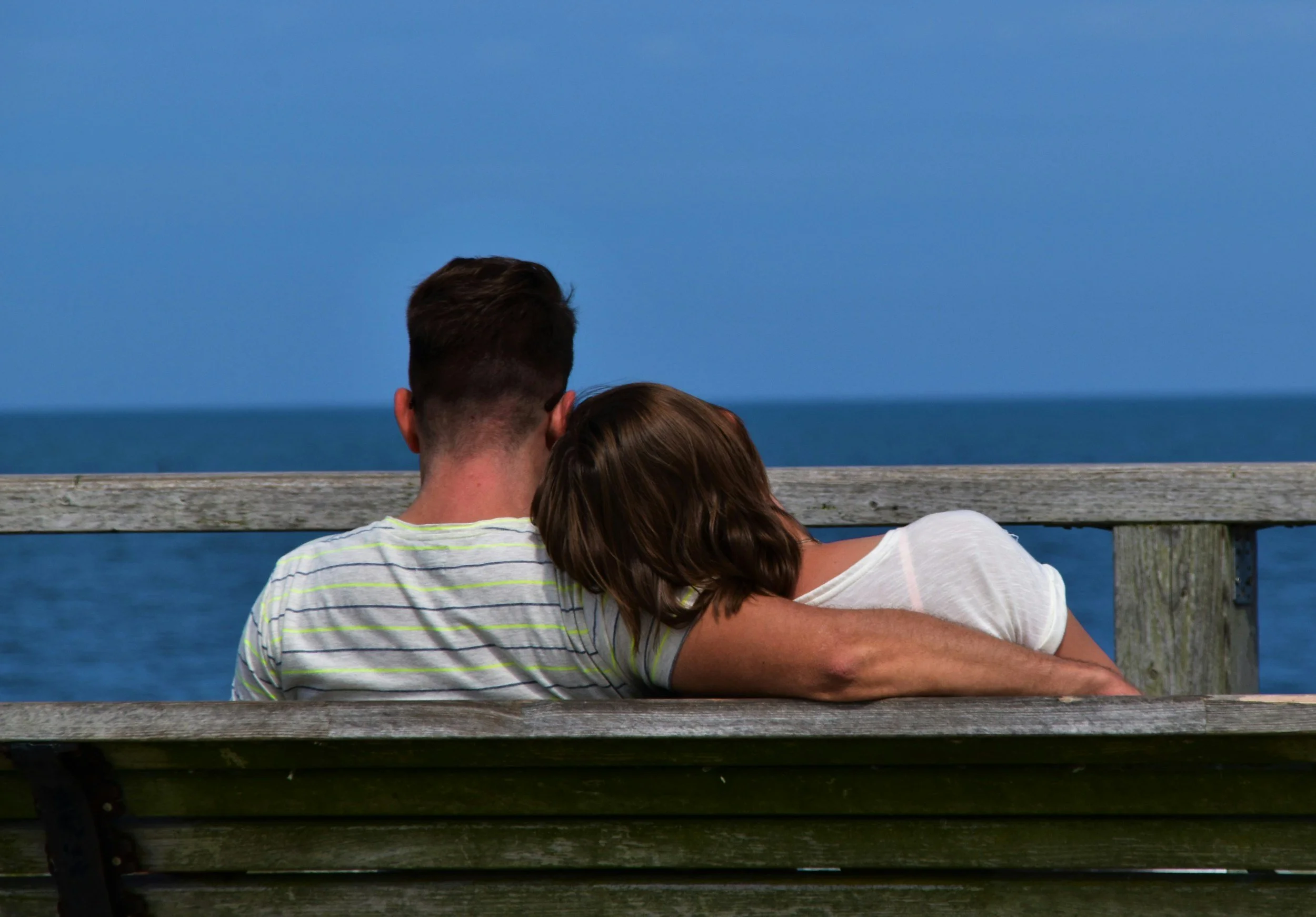 A couple sitting on a wooden bench by the ocean, with the woman resting her head on the man's shoulder, facing the water and horizon.