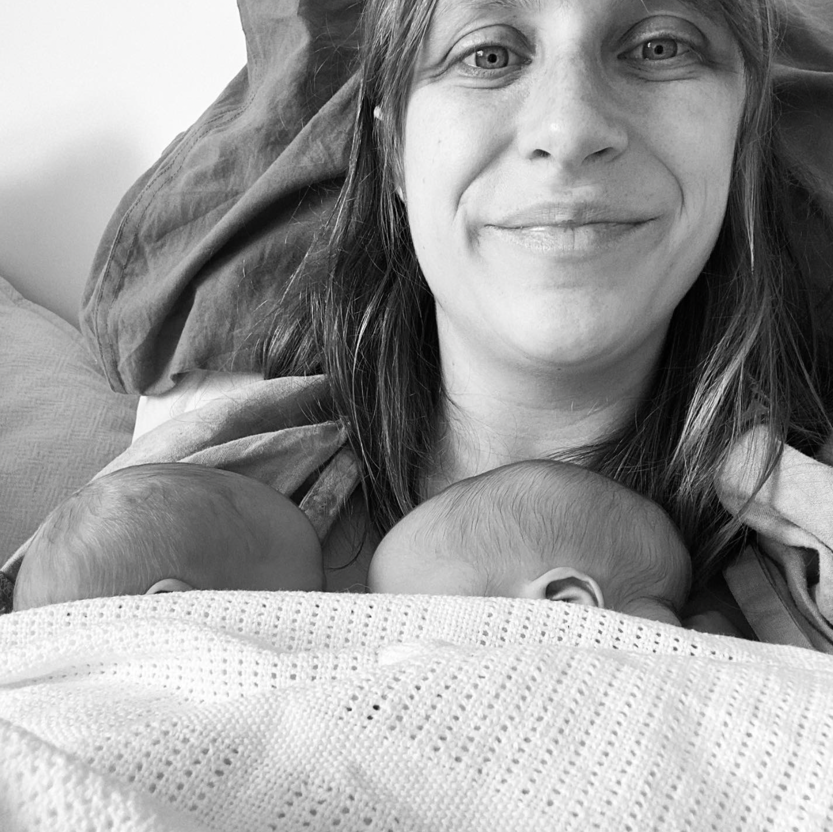 A black and white photo of a woman smiling, holding newborn twins close to her chest enjoying skin-to-skin with their heads resting on her.