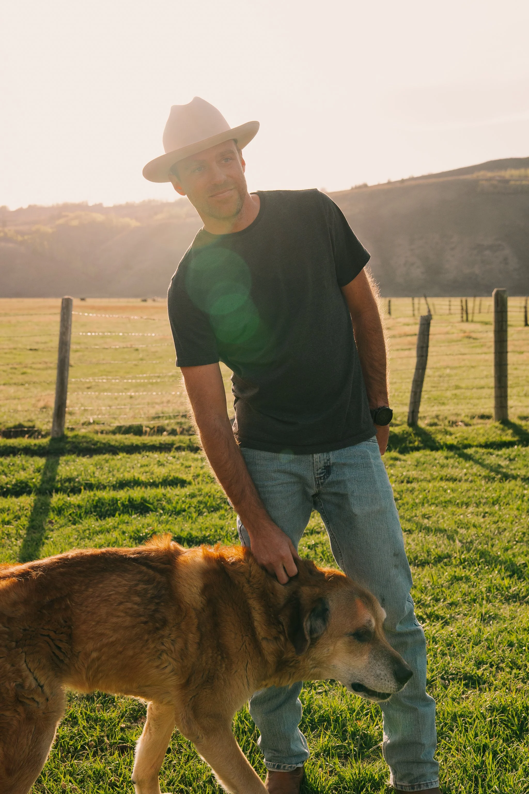 A man in a black t-shirt, light jeans, and a beige hat standing in a grassy field with a large dog, possibly a golden retriever, during sunset with hills in the background.