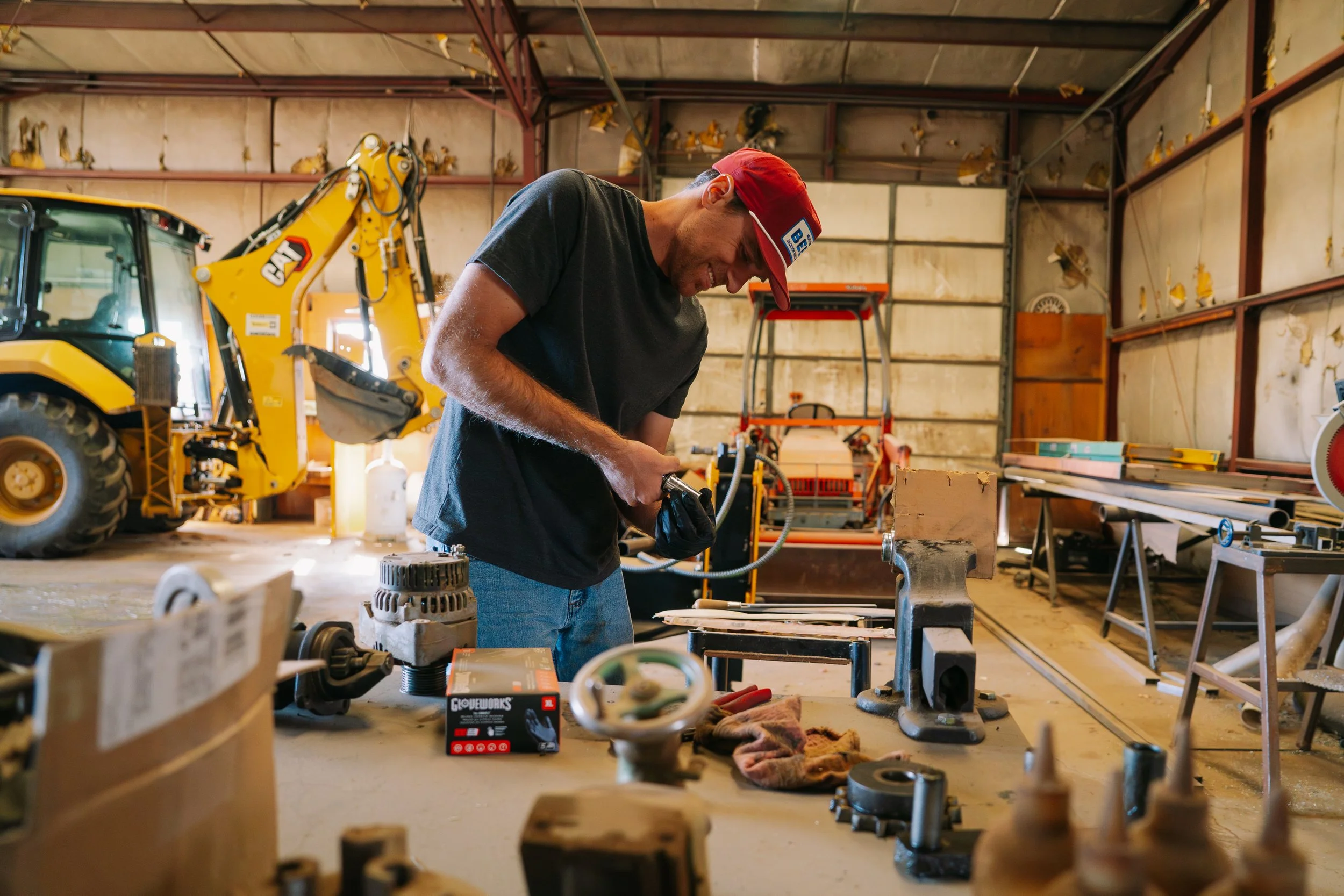 A man wearing a red cap working on a project in a workshop filled with tools and equipment, including a yellow backhoe and various machinery.