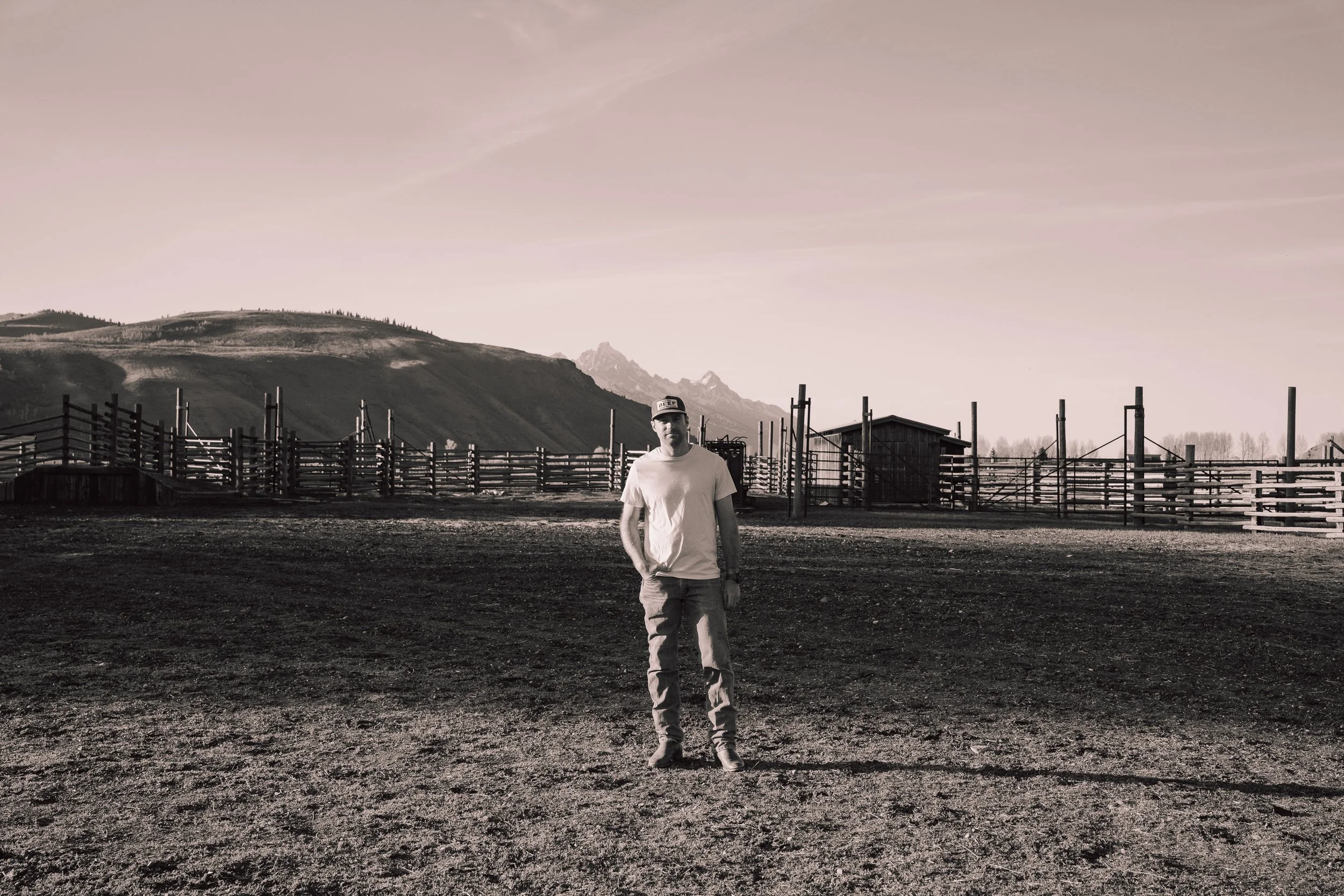 A man standing outdoors on a dirt surface with mountains and a wooden fence in the background, under a cloudy sky.