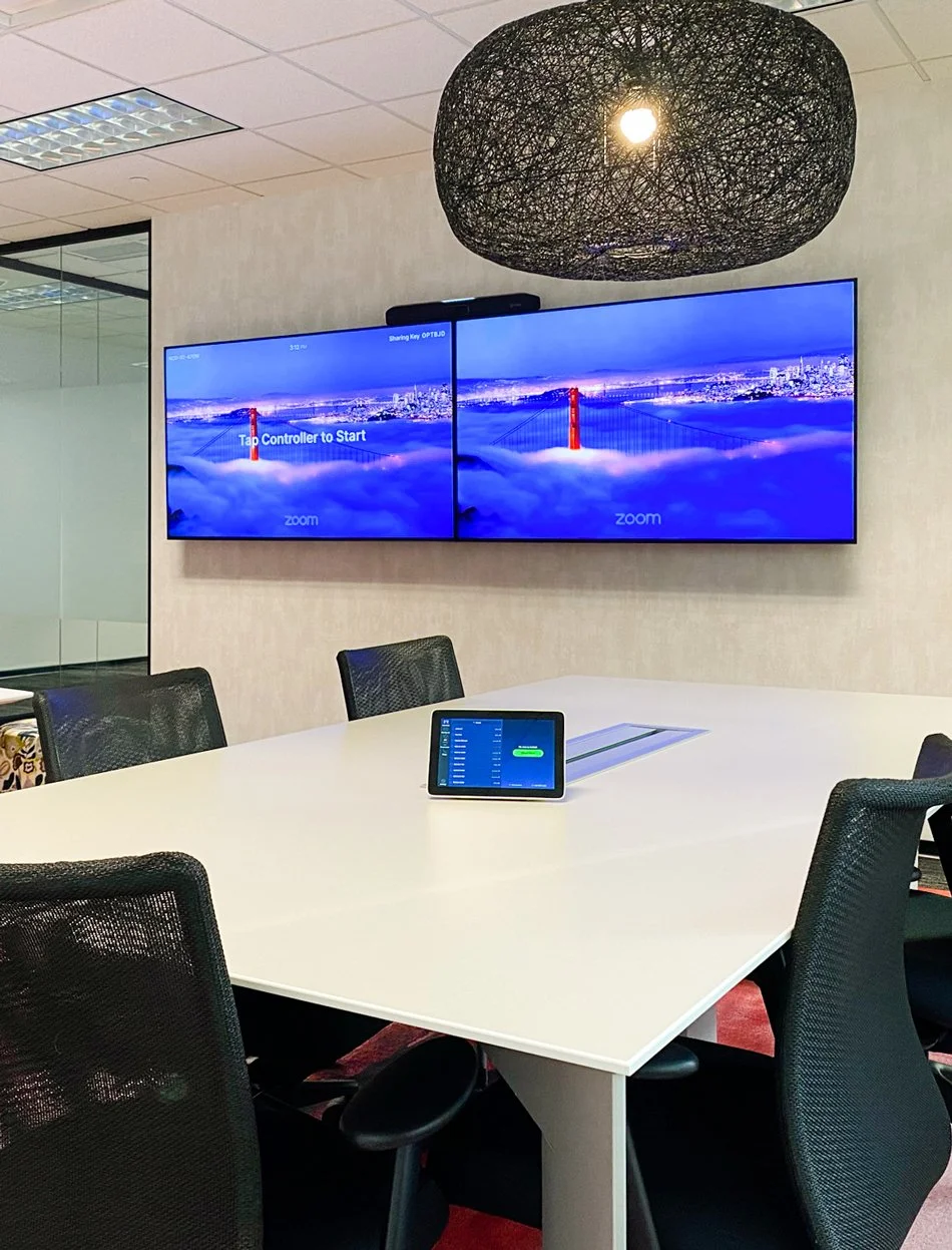 Modern conference room featuring dual video displays, a tabletop touchscreen control panel, and integrated audiovisual conferencing technology.
