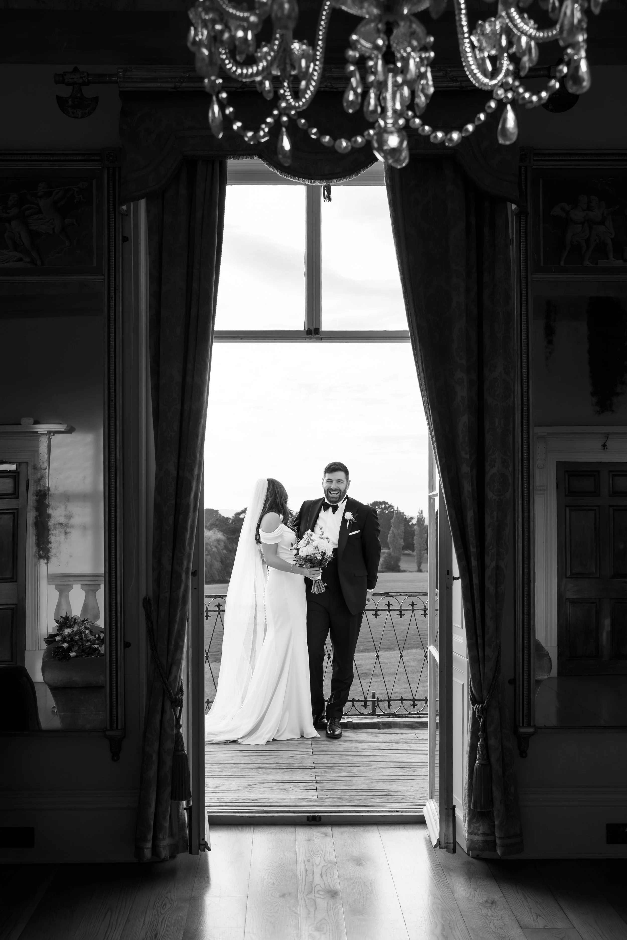 A bride and groom stand on a balcony, framed by open double doors with ornate curtains and a chandelier hanging above. The bride wears a white gown with a veil and holds a bouquet, while the groom wears a tuxedo and smiles at her. Outside, scenic tre
