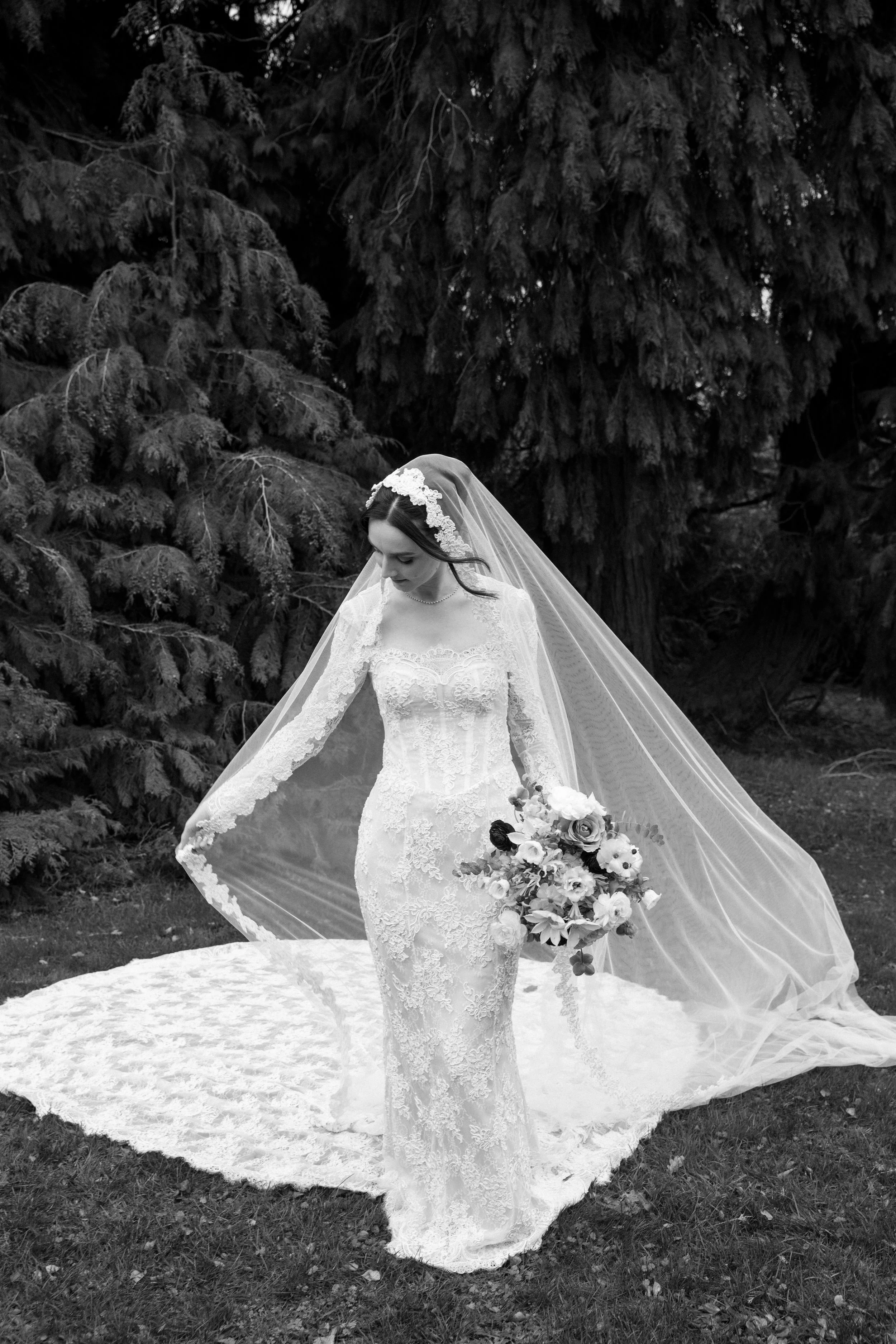 A bride in a lace wedding gown and veil holding a bouquet of flowers standing outdoors on grass with large pine trees in the background.