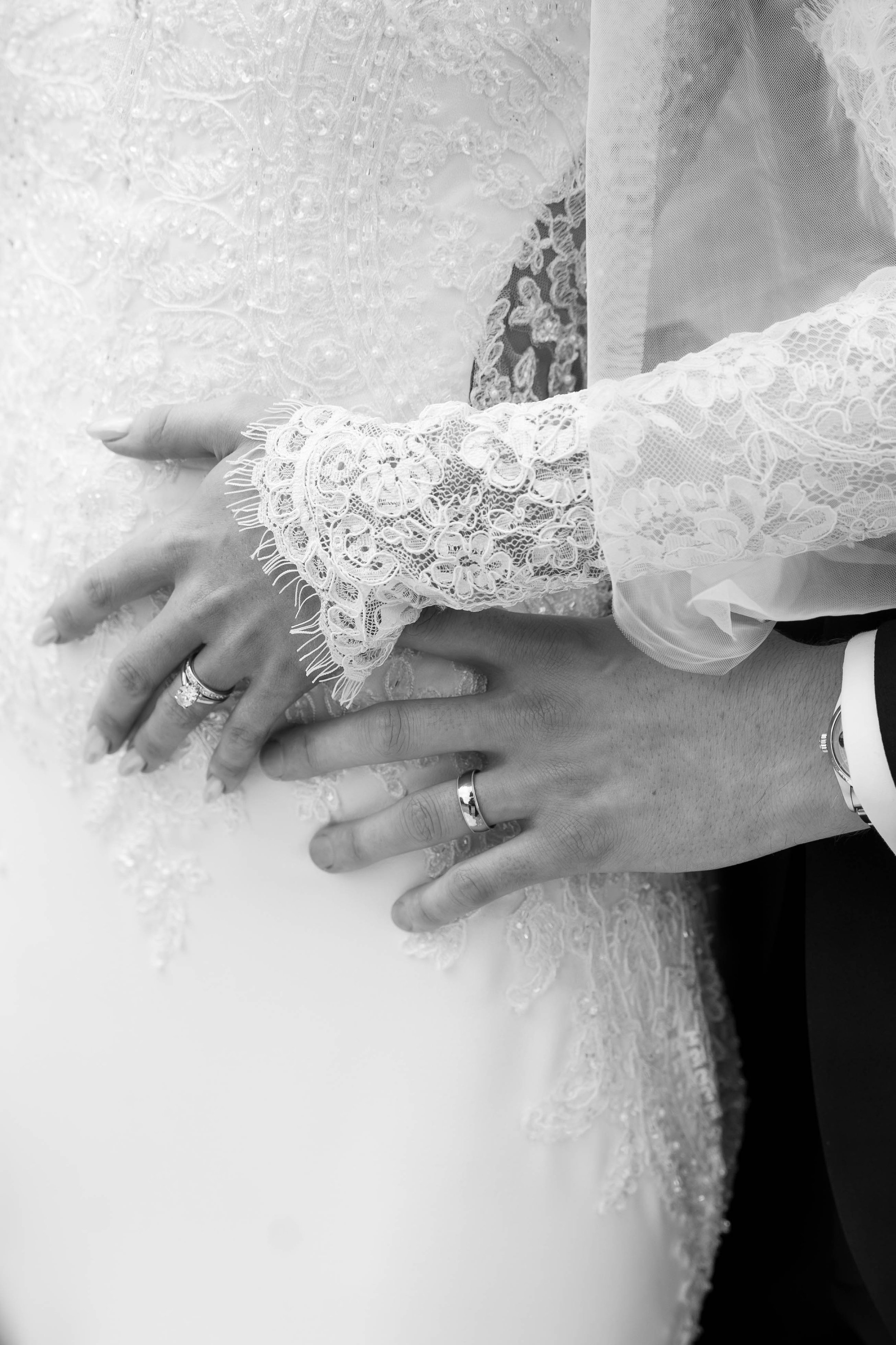Close-up of hands with wedding rings resting on a lace wedding dress, detailed lace fabric visible on the dress.