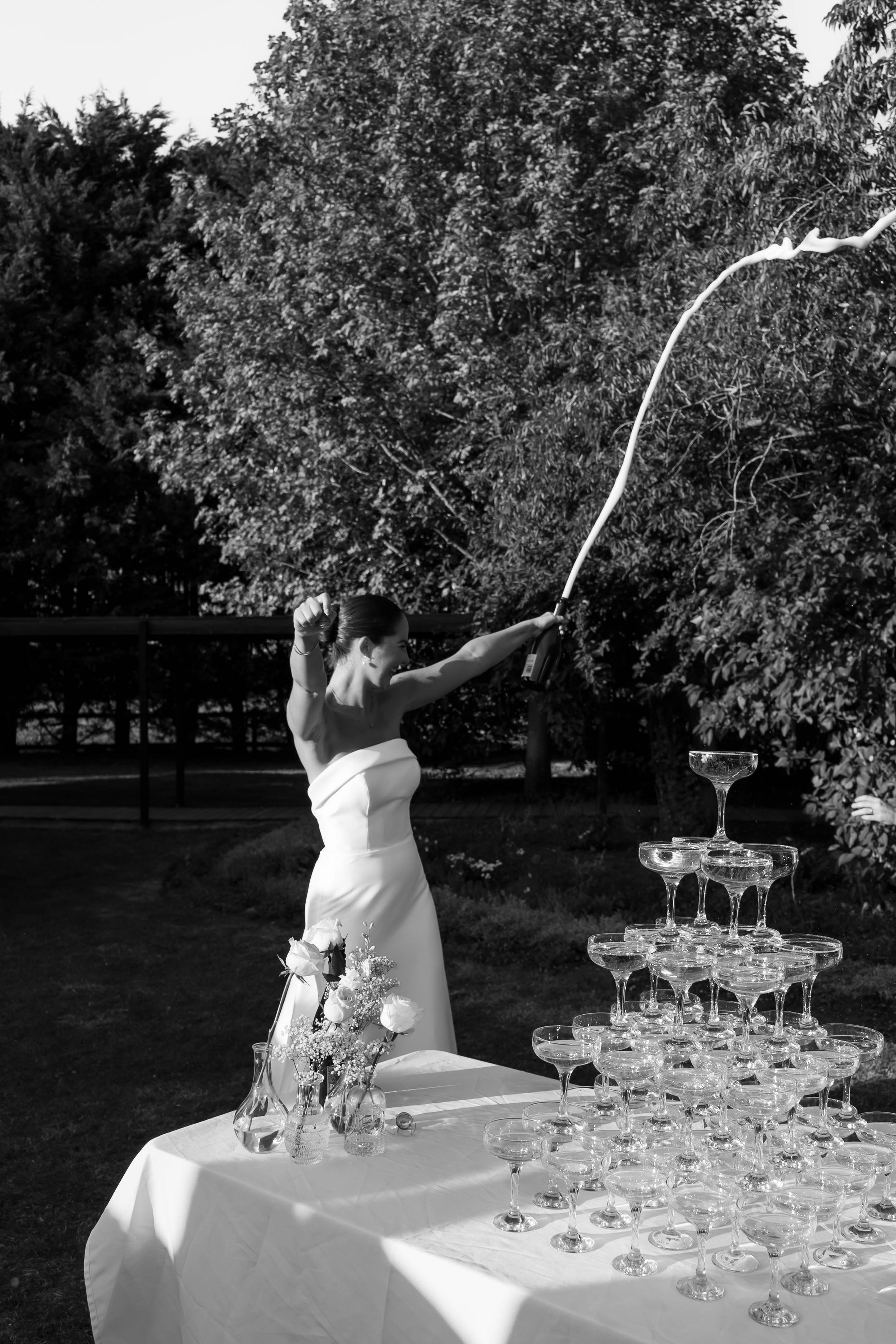 A woman in a white dress is celebrating by knocking over a champagne tower at an outdoor event. She is smiling and holding a champagne bottle. There is a champagne bouquet on the table and a stack of coupe glasses arranged in a pyramid. Trees are vis