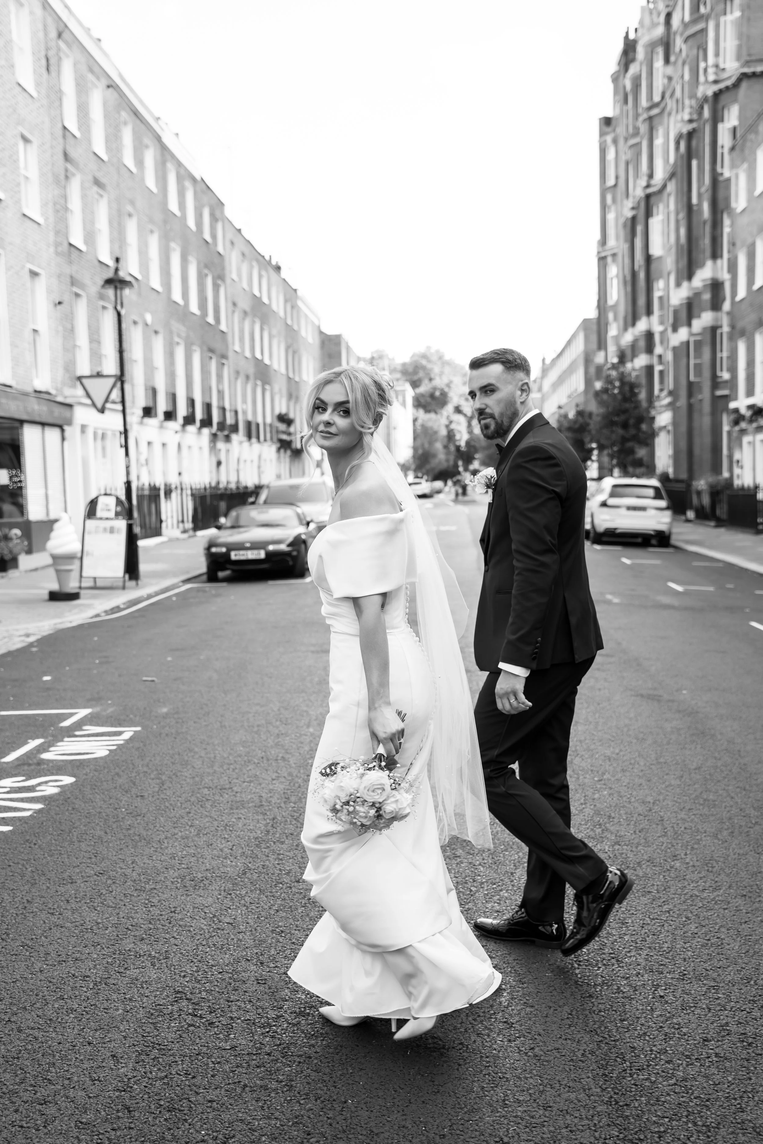 A bride in a white wedding gown and a groom in a black tuxedo walk on a city street, carrying a bouquet of flowers and facing opposite directions.