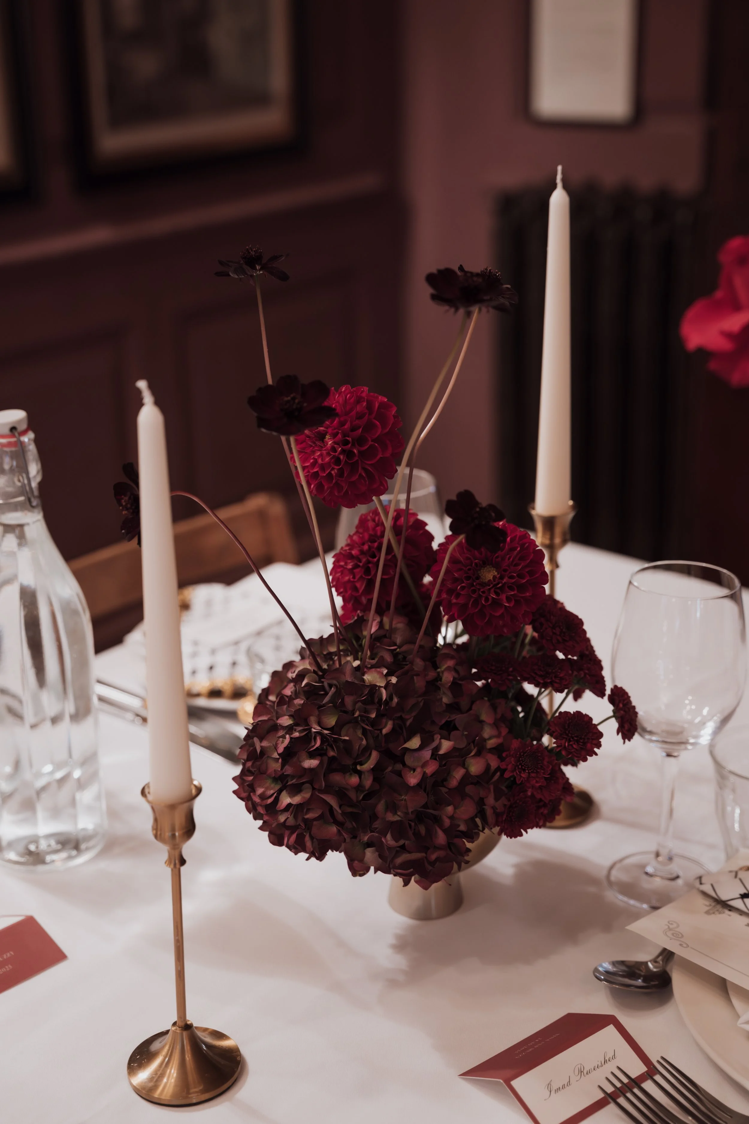 A centerpiece with dark red and purple flowers on a dining table, flanked by tall white candles in gold holders. The table is set with glassware, a bottle of water, and place cards. The background has framed pictures on a dark wall.