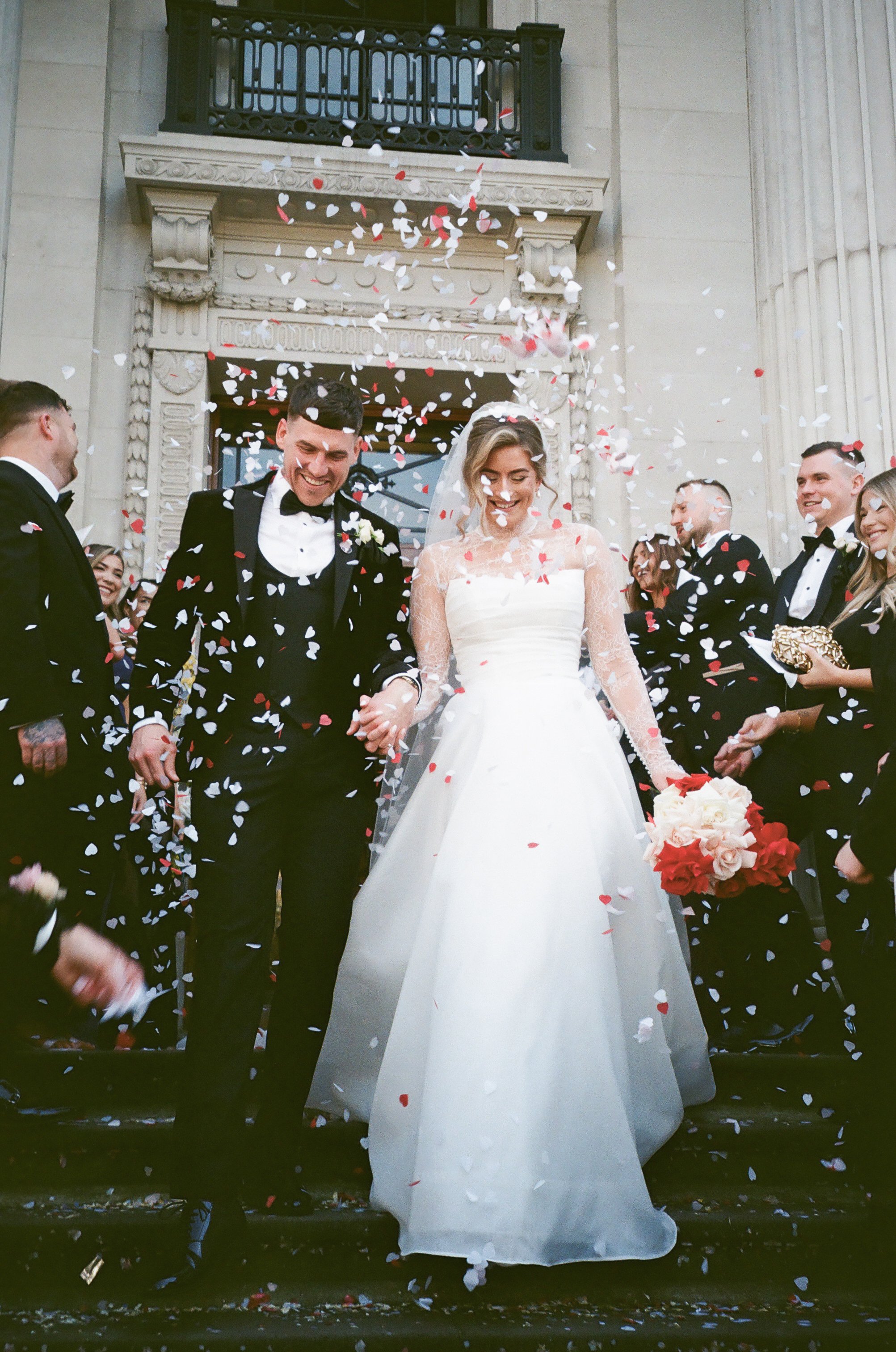 A bride and groom holding hands and walking down stairs, surrounded by friends and family, during a wedding celebration with confetti falling around them.