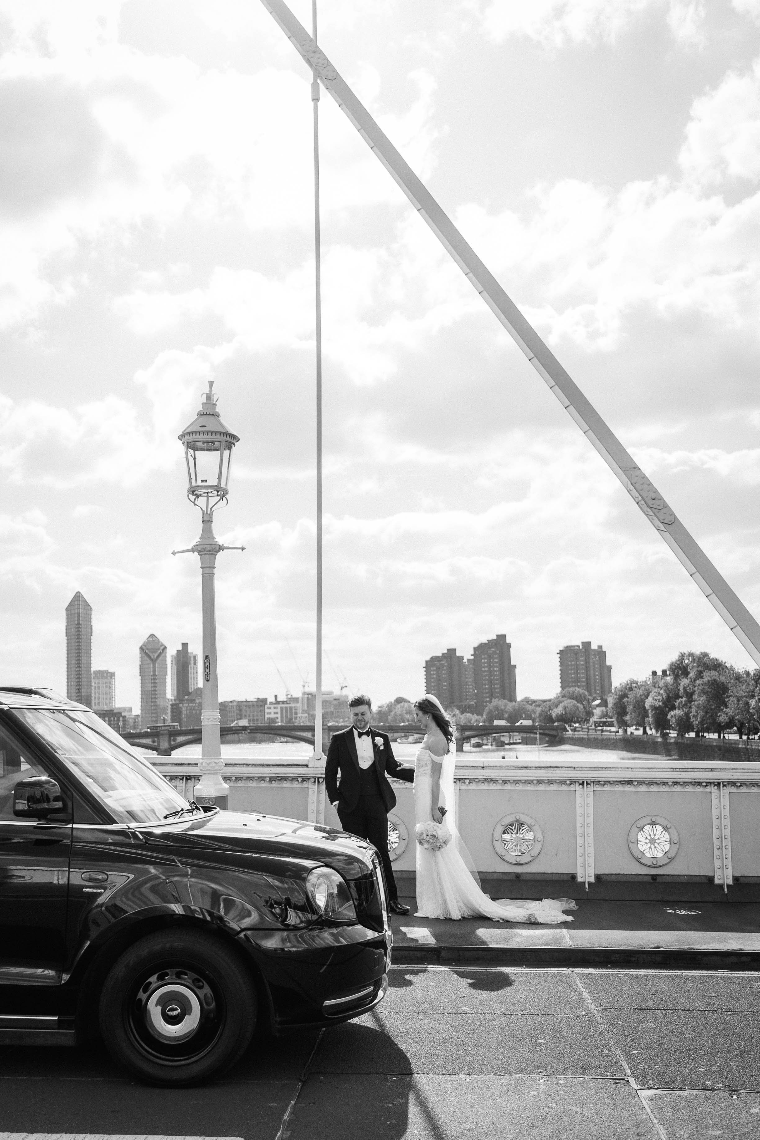 A black and white photo of a bride and groom standing on a bridge with a cityscape in the background. The bride is in a long white wedding dress holding a bouquet, and the groom is in a black tuxedo.