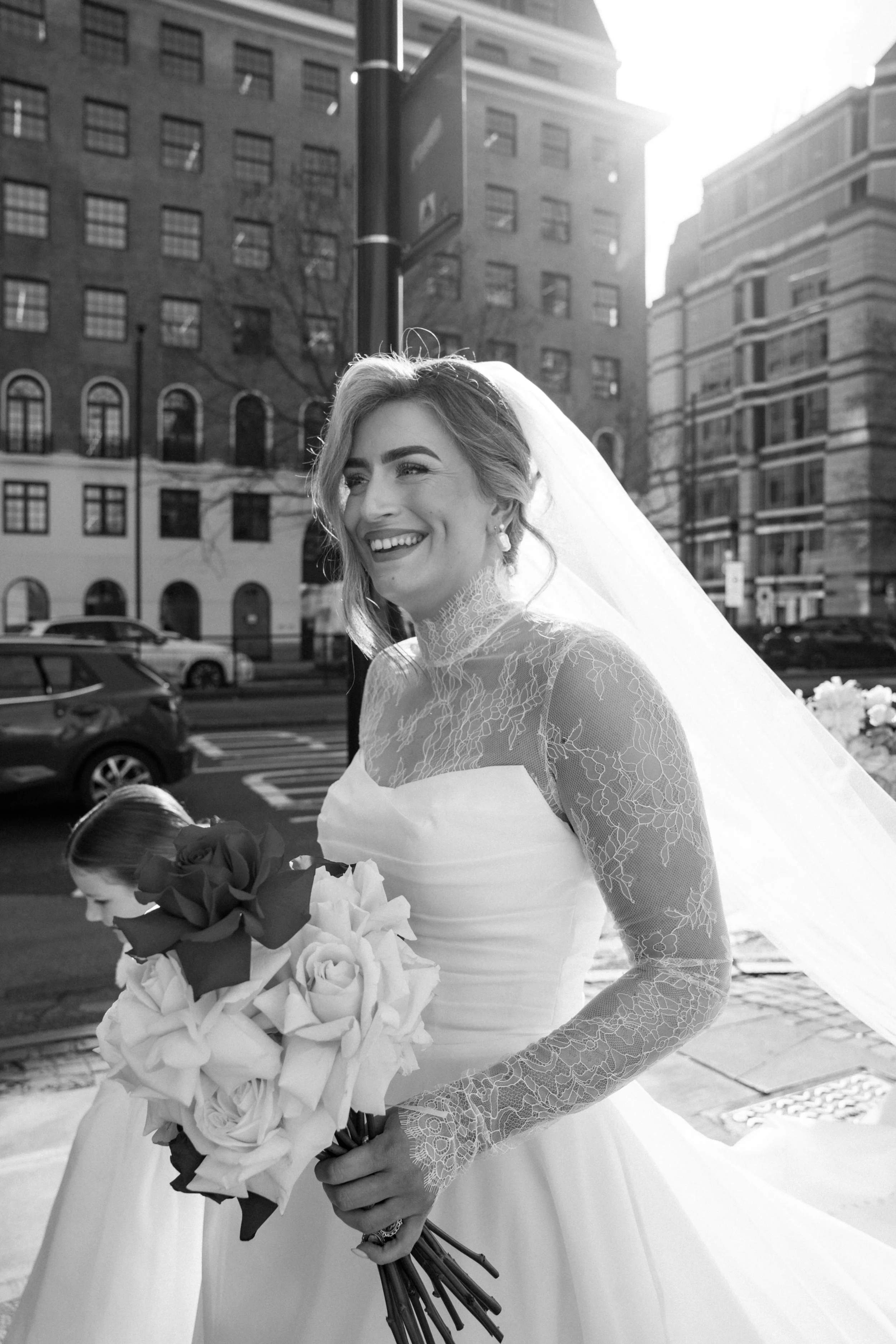 Woman in a wedding dress holding a bouquet of flowers, standing outdoors in front of city buildings.