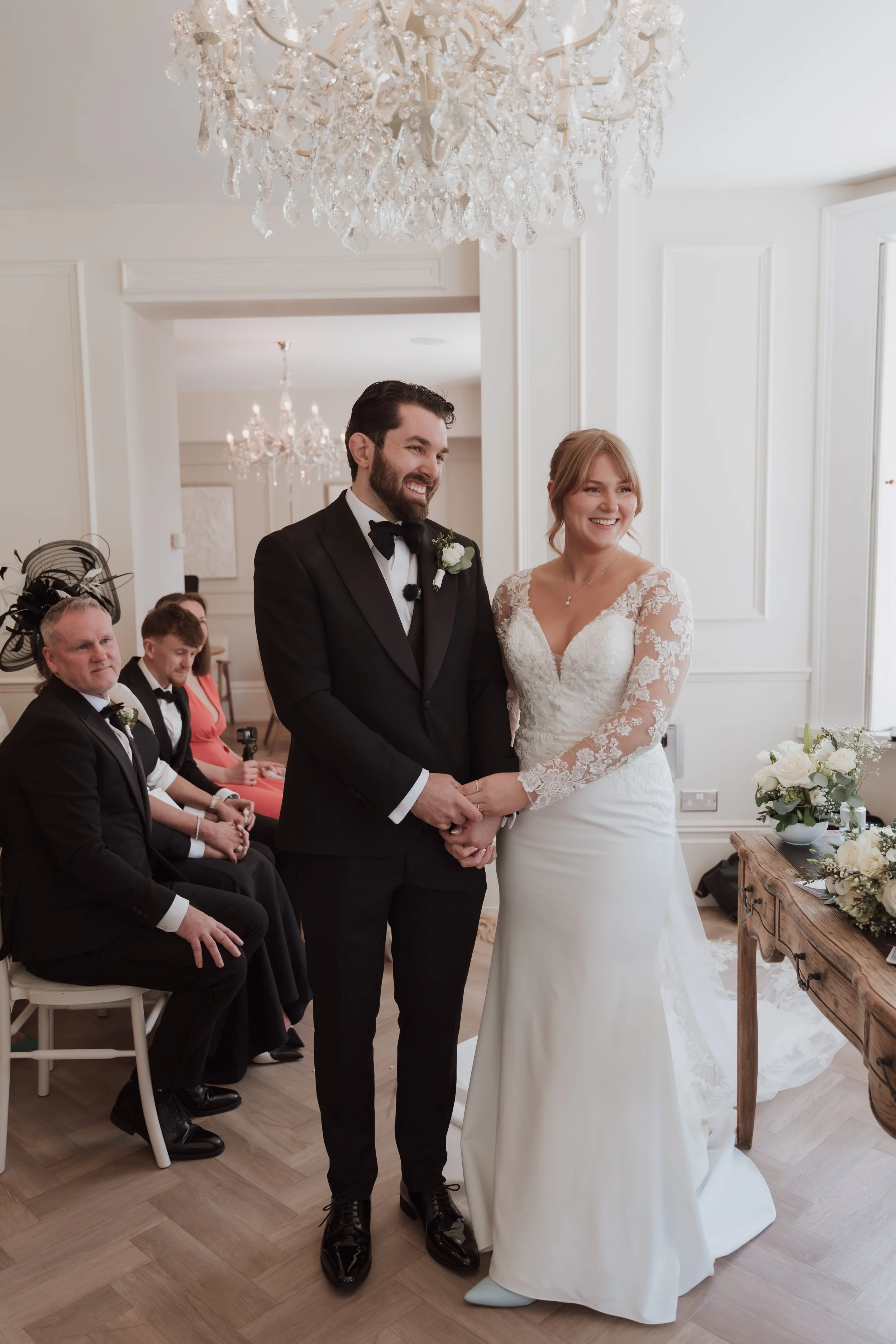 A bride and groom holding hands during their wedding ceremony in an elegant, well-lit room with chandeliers and white walls. The bride is wearing a lace wedding dress, and the groom is dressed in a black tuxedo with a bow tie. Several seated guests i