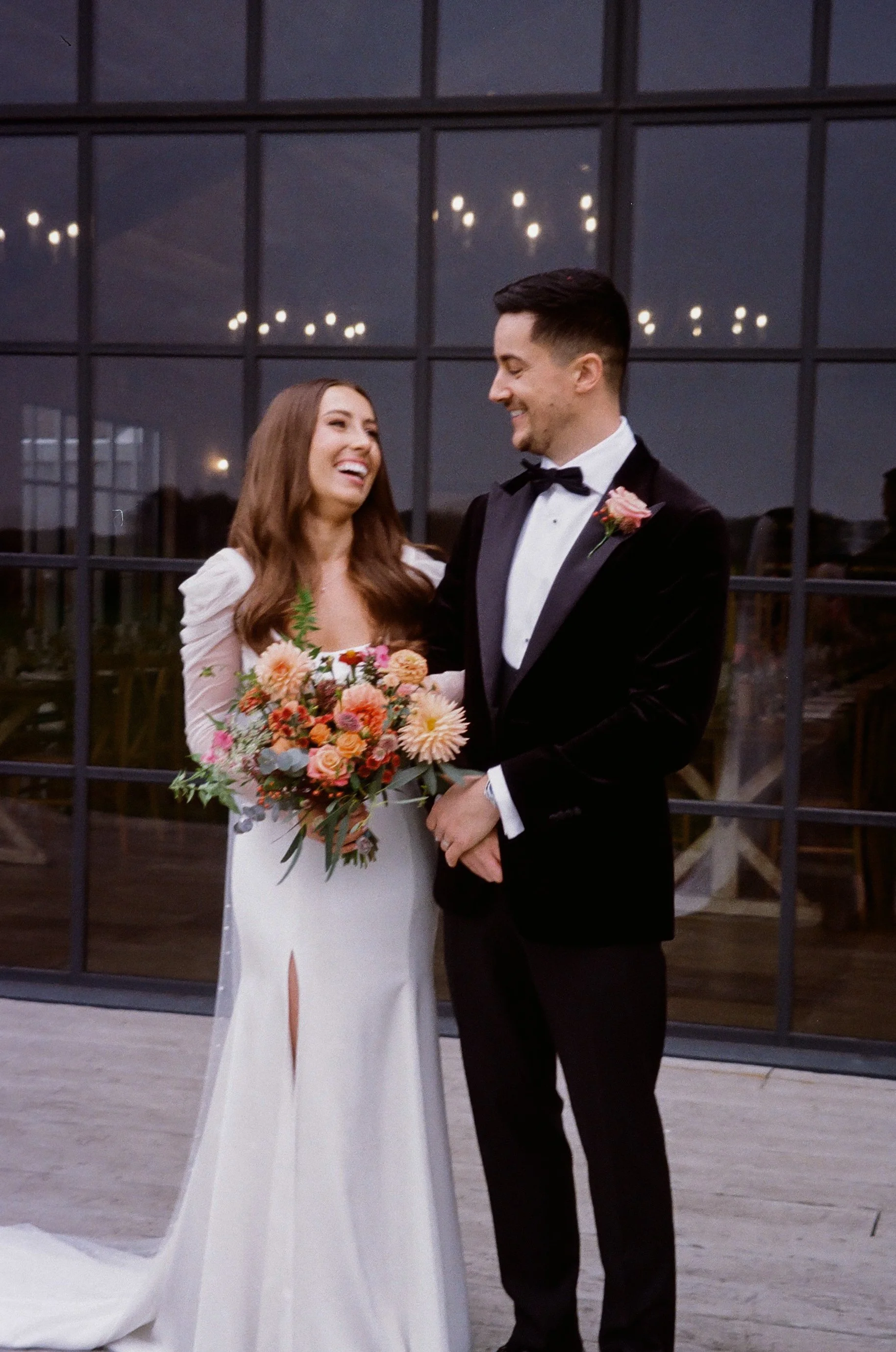 A bride and groom smiling at each other during their wedding, standing inside a modern building with large glass windows reflecting ceiling lights. The bride holds a colorful bouquet while the groom wears a black tuxedo with a pink flower boutonniere.