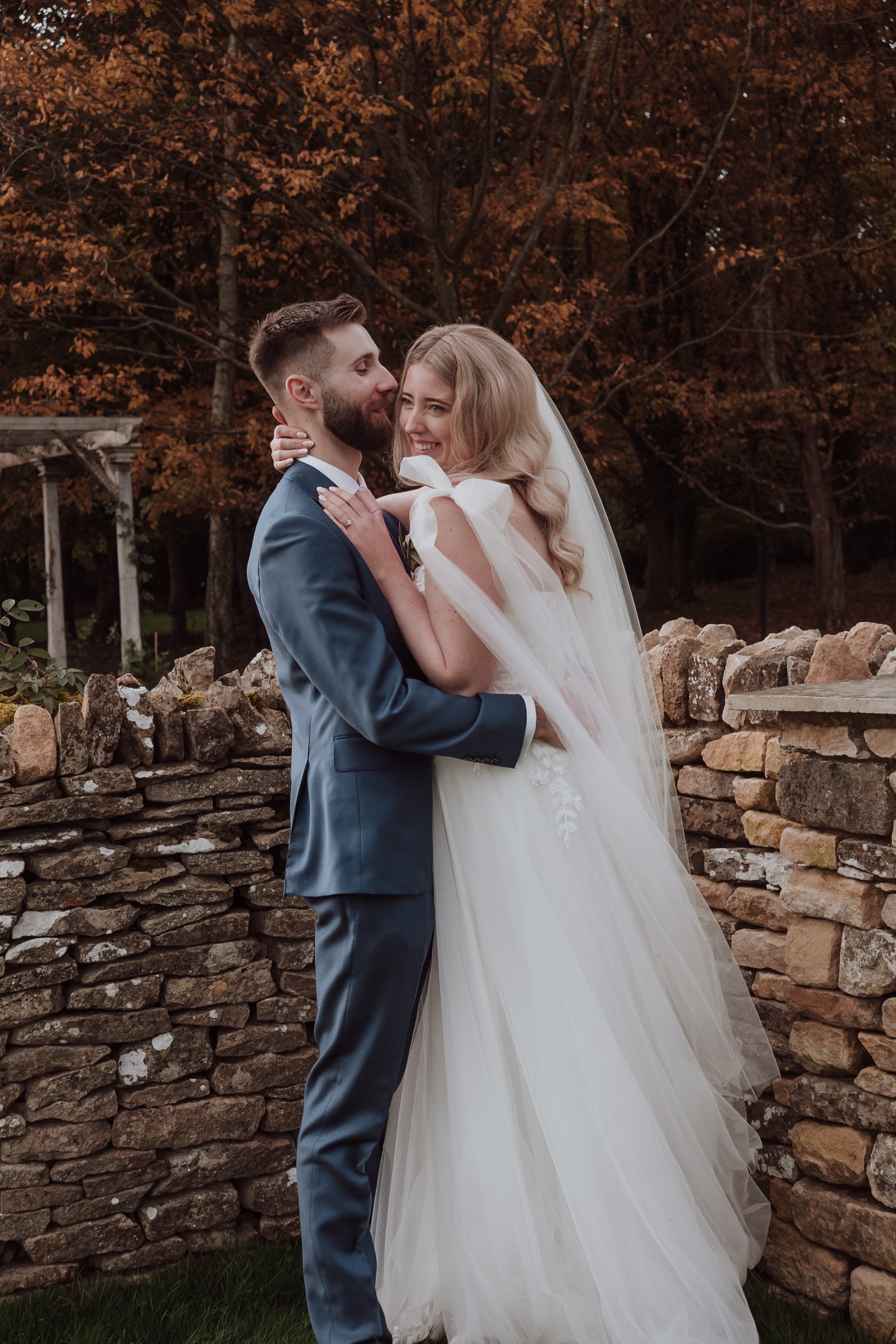 A bride and groom sharing a joyful moment outdoors during their wedding, standing in front of a stone wall with autumn trees in the background.