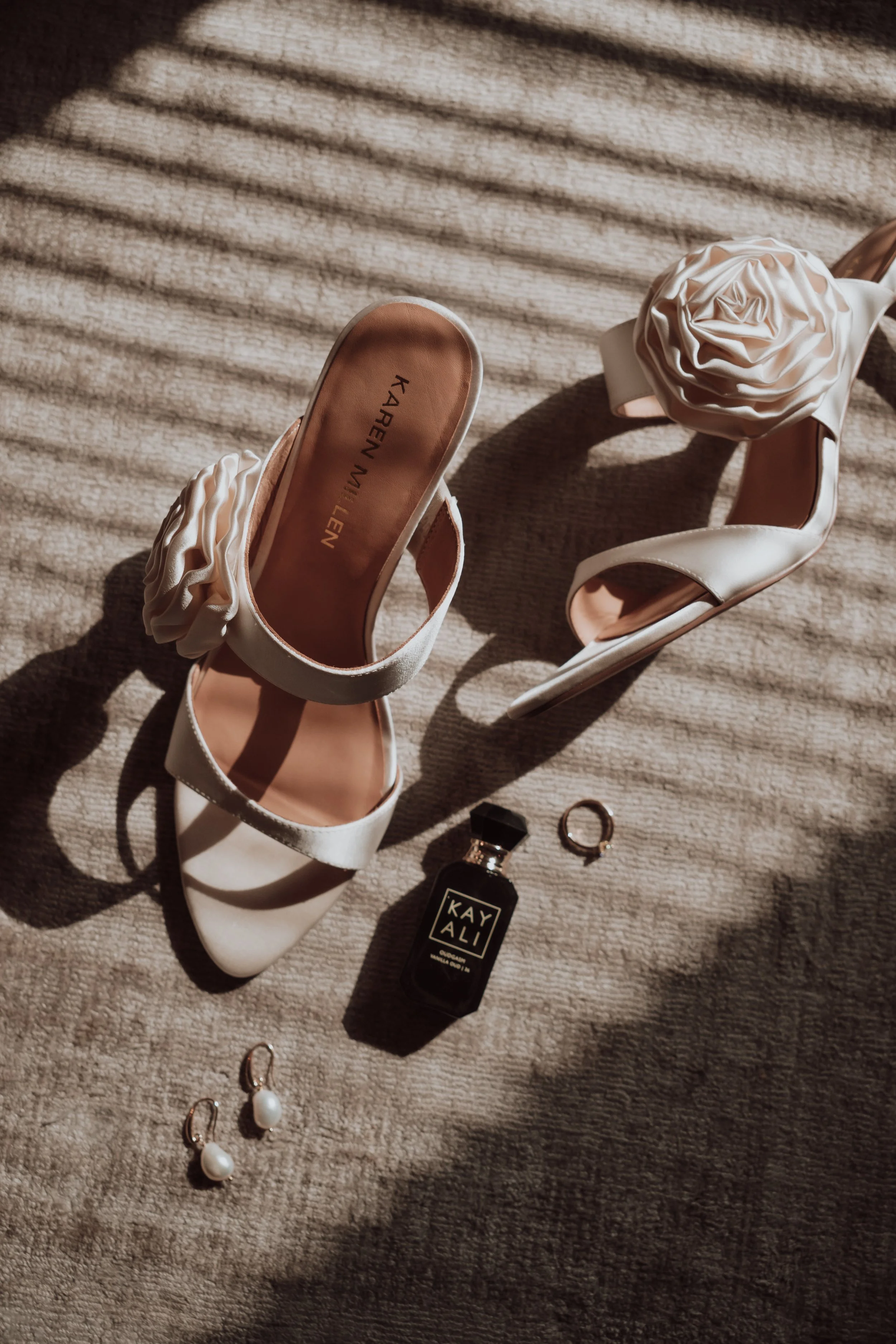 White high-heeled shoes with large fabric flower decorations, jewelry including earrings and rings, a perfume bottle labeled "KAYALI", and a small nail polish bottle, all arranged on a textured beige carpet with shadows from window blinds.