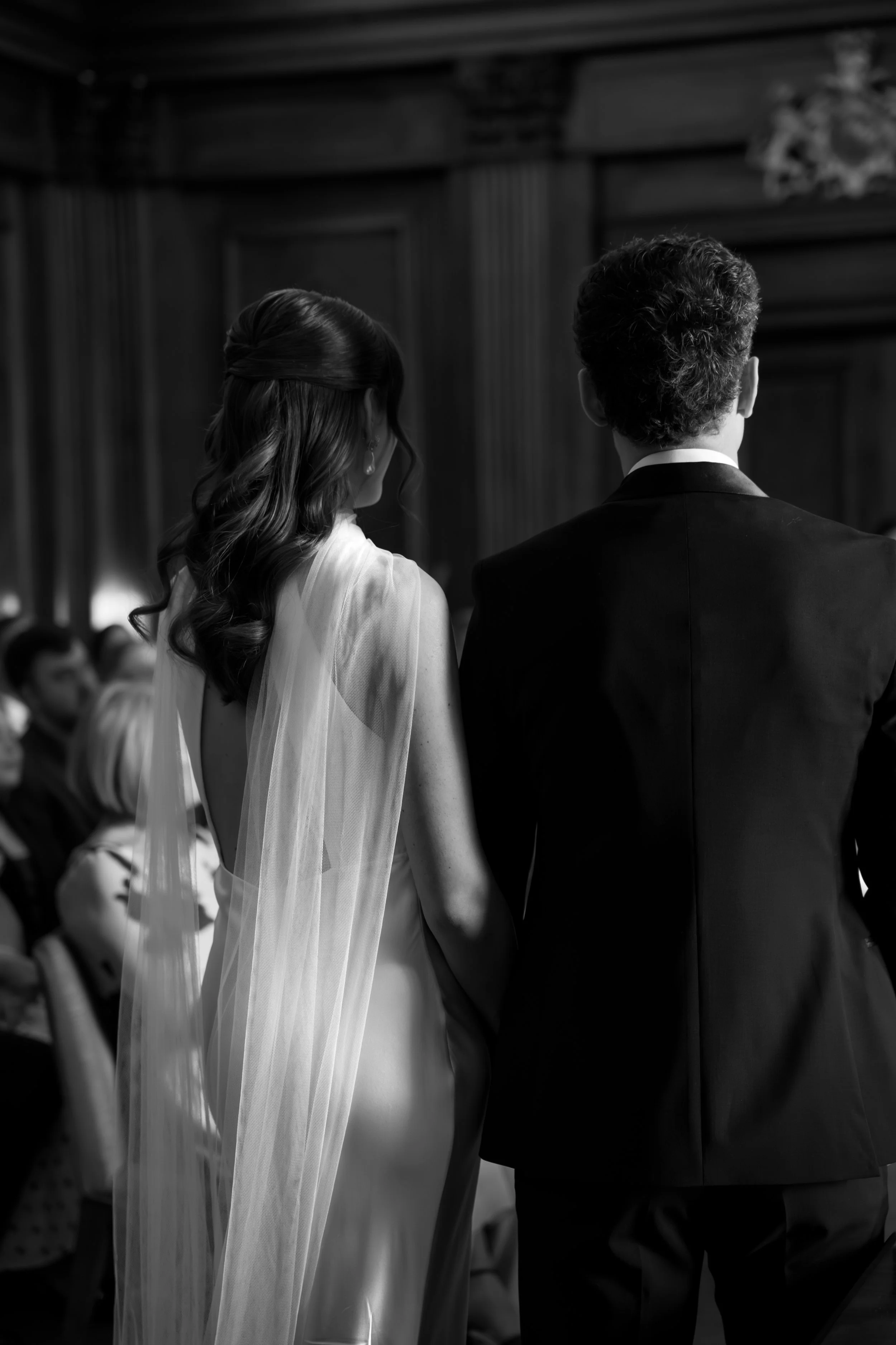 Black and white photo of a bride and groom standing side by side, facing away from the camera during their wedding ceremony, with guests seated in the background.