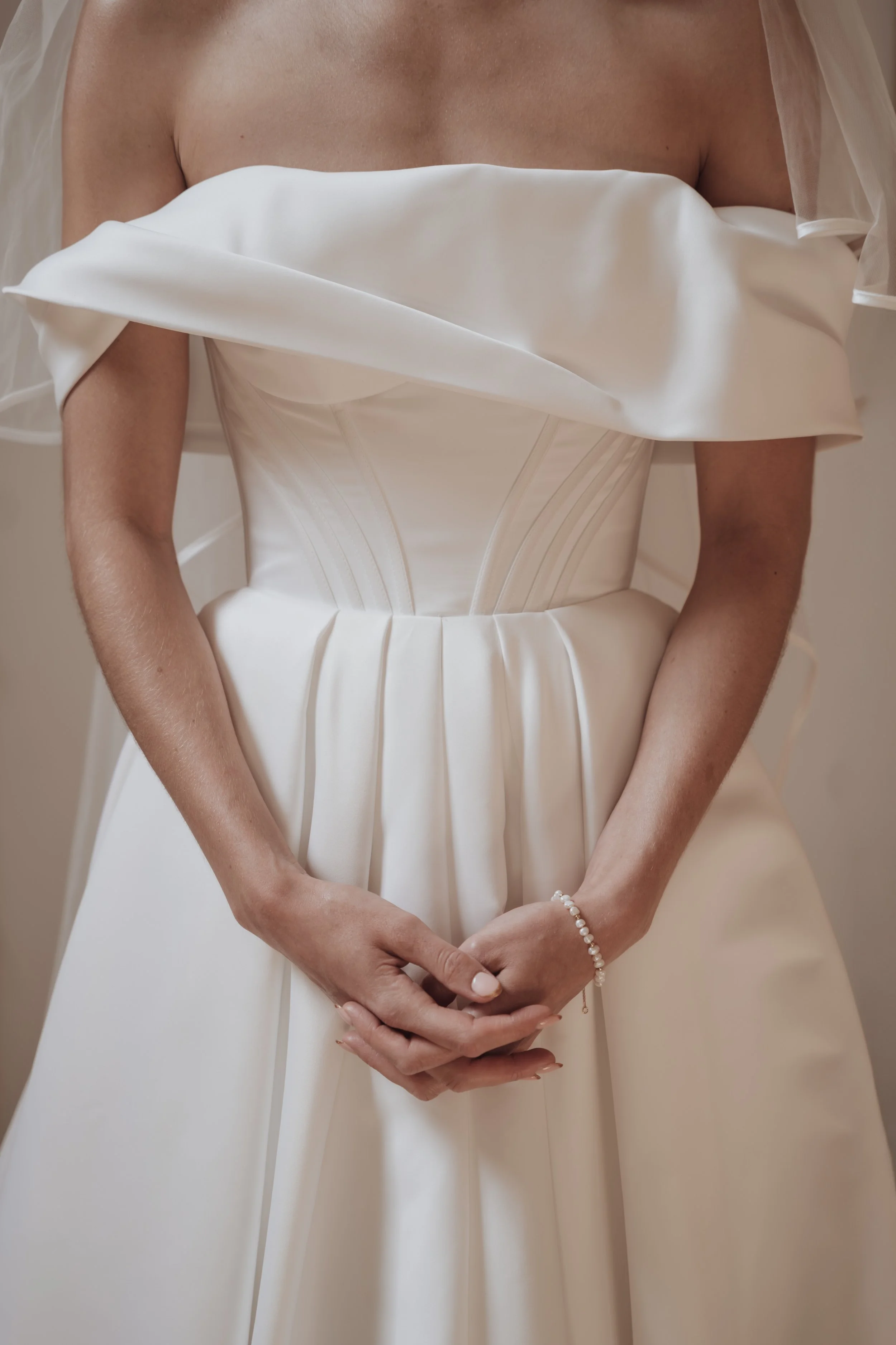 Close-up of a bride in a white wedding dress with off-the-shoulder sleeves and pleated details, holding her hands in front of her with a pearl bracelet on her wrist.
