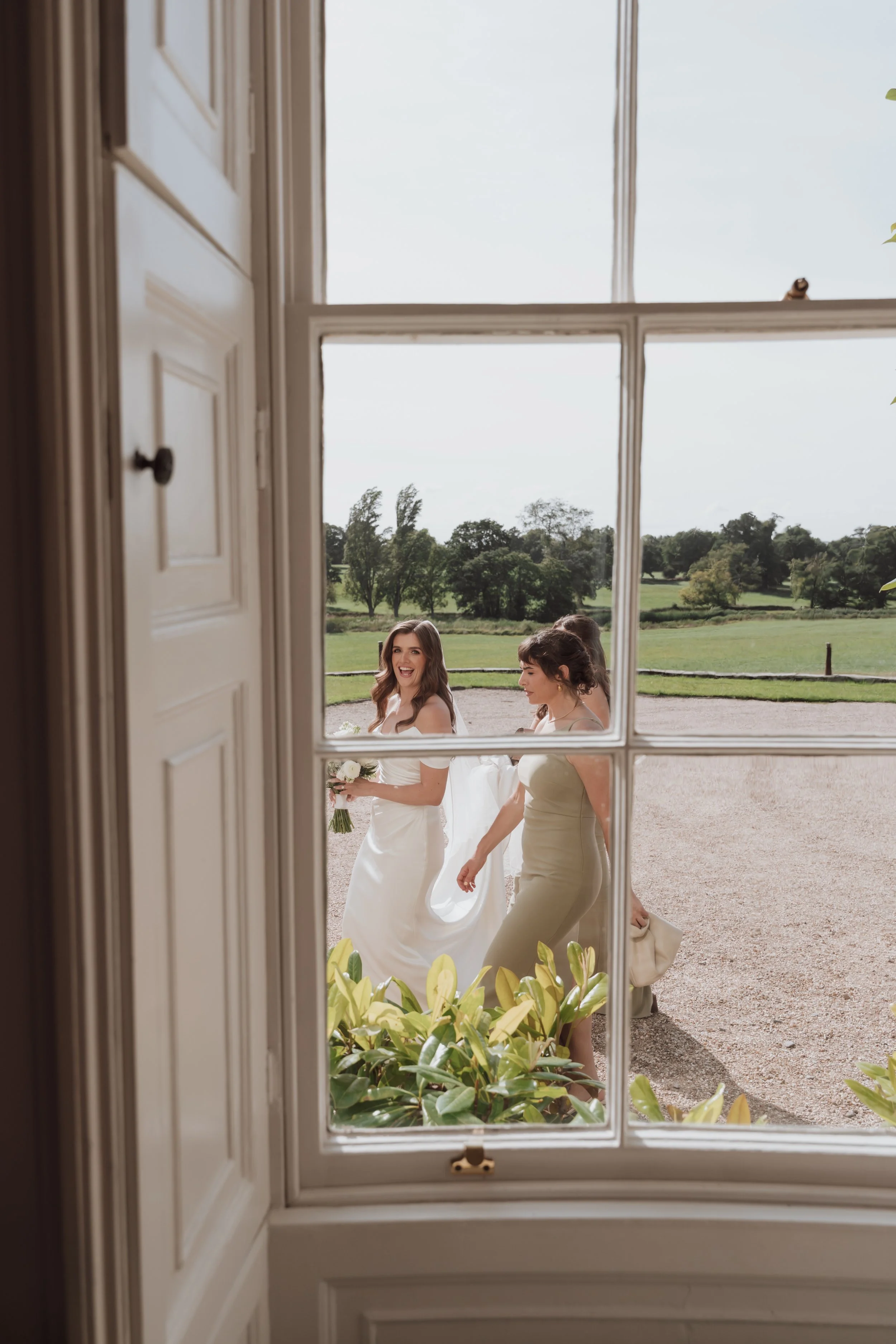 Photo of three women in elegant dresses walking outside through a large window, with a green landscape and trees in the background.
