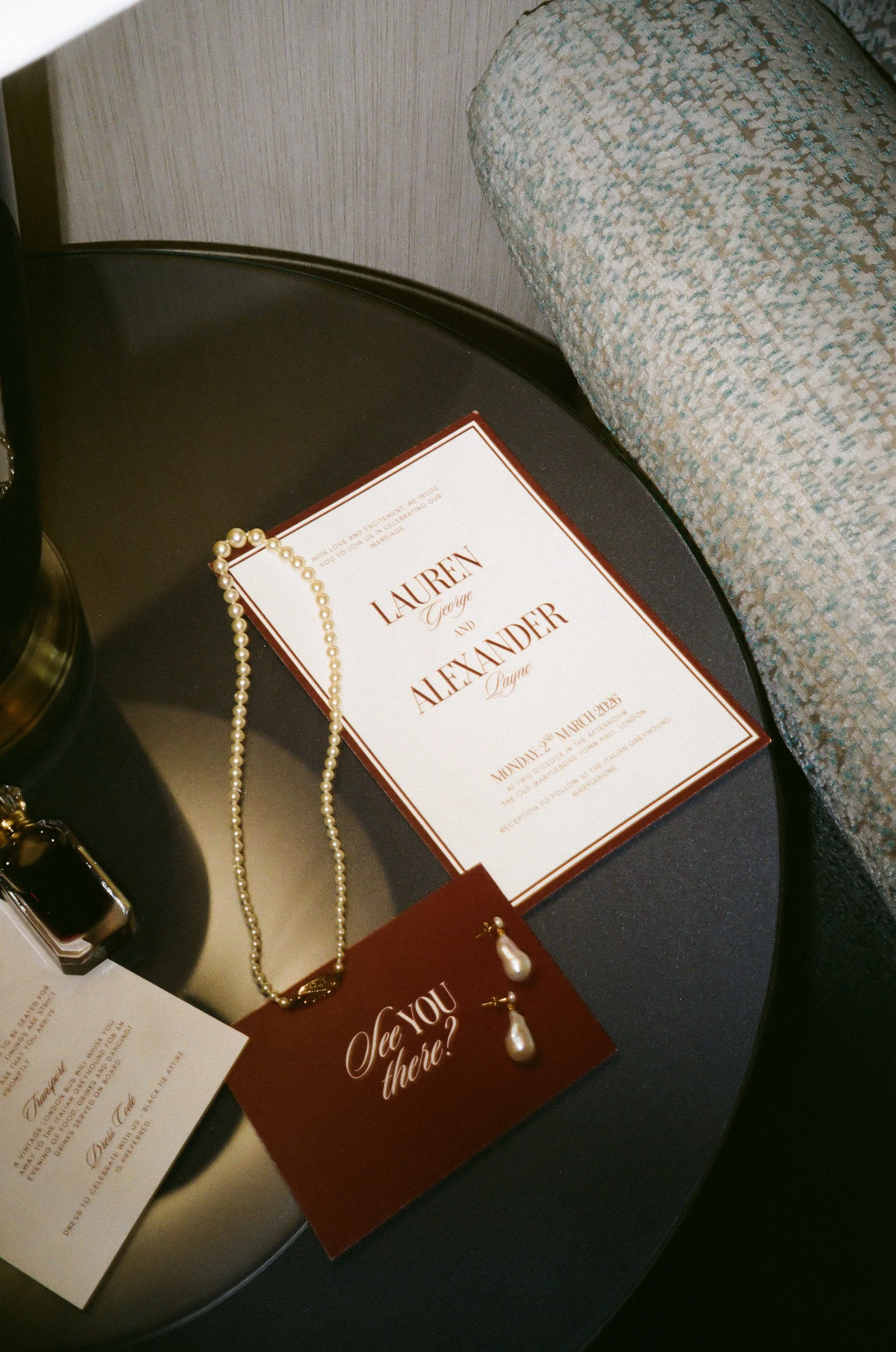 Wedding invitation card with names Lauren George and Alexander, wedding date March 21, 2025, on a black round table alongside pearl jewelry, a small bottle of perfume, and a card with the text "See you there?" on a burgundy background.