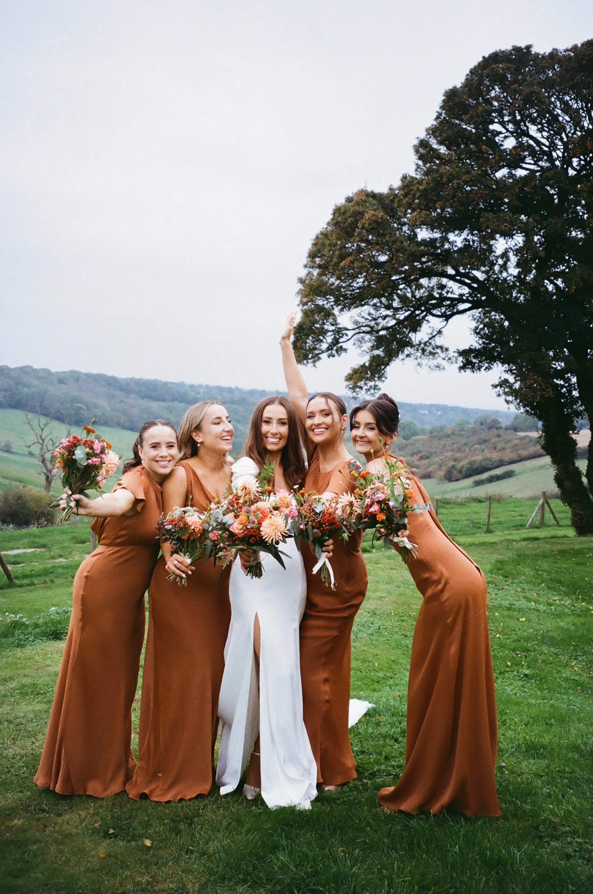 Group of women, dressed in a mix of white and rust-colored dresses, celebrating outdoors with bouquets of flowers, smiling and posing for a photo in a green field with trees and hills in the background.
