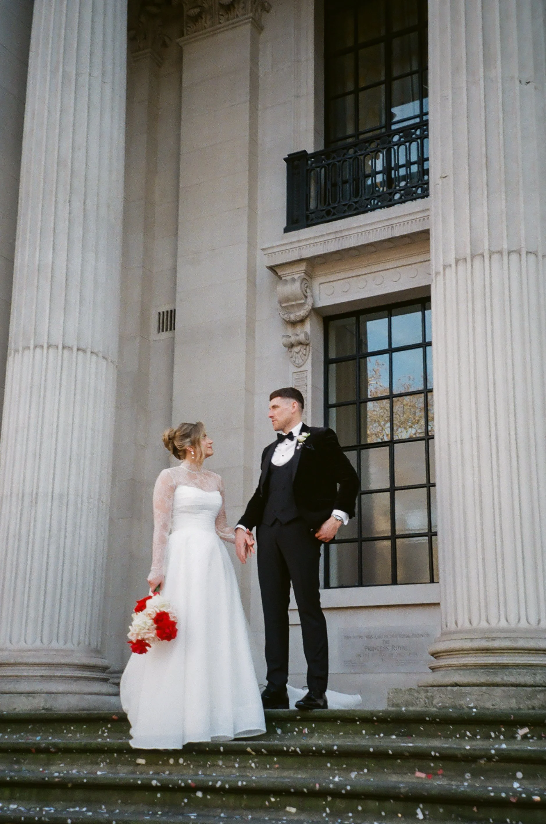 A bride and groom standing on the steps of a neoclassical building with large columns, looking at each other. The bride is holding a bouquet of red and white flowers, and the groom is dressed in a black tuxedo with a bow tie.