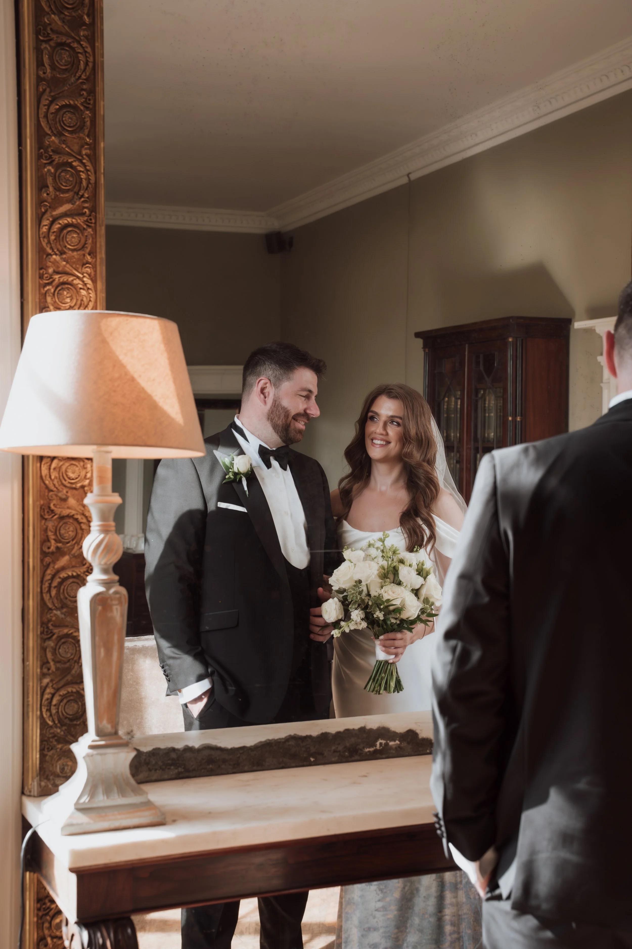 A man and a woman dressed in wedding attire smiling at each other in front of a mirror, with a person in a tuxedo partially visible in the foreground.