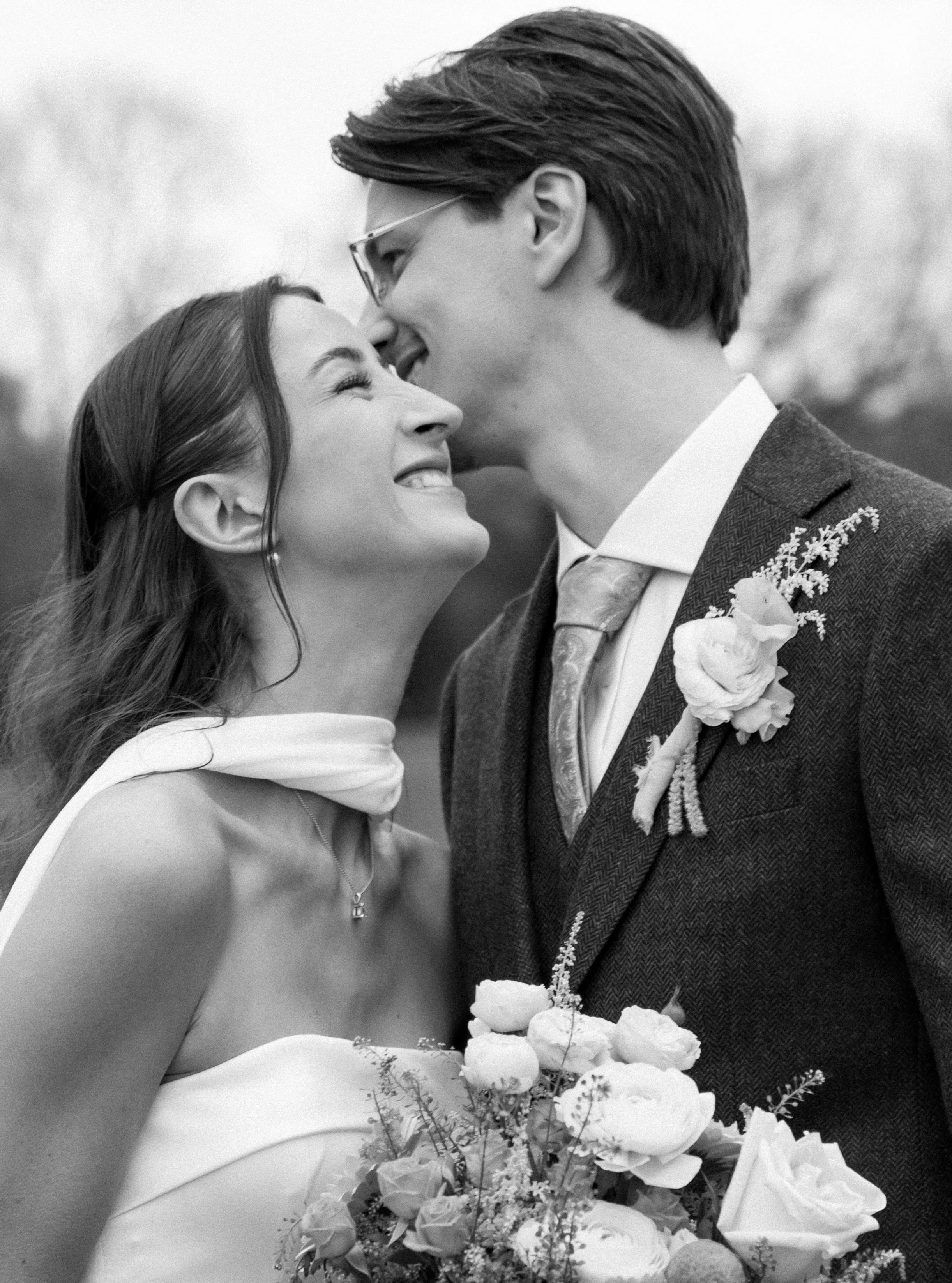 Black and white photograph of a couple at their wedding, with the bride smiling and holding a bouquet, and the groom leaning in for a kiss.