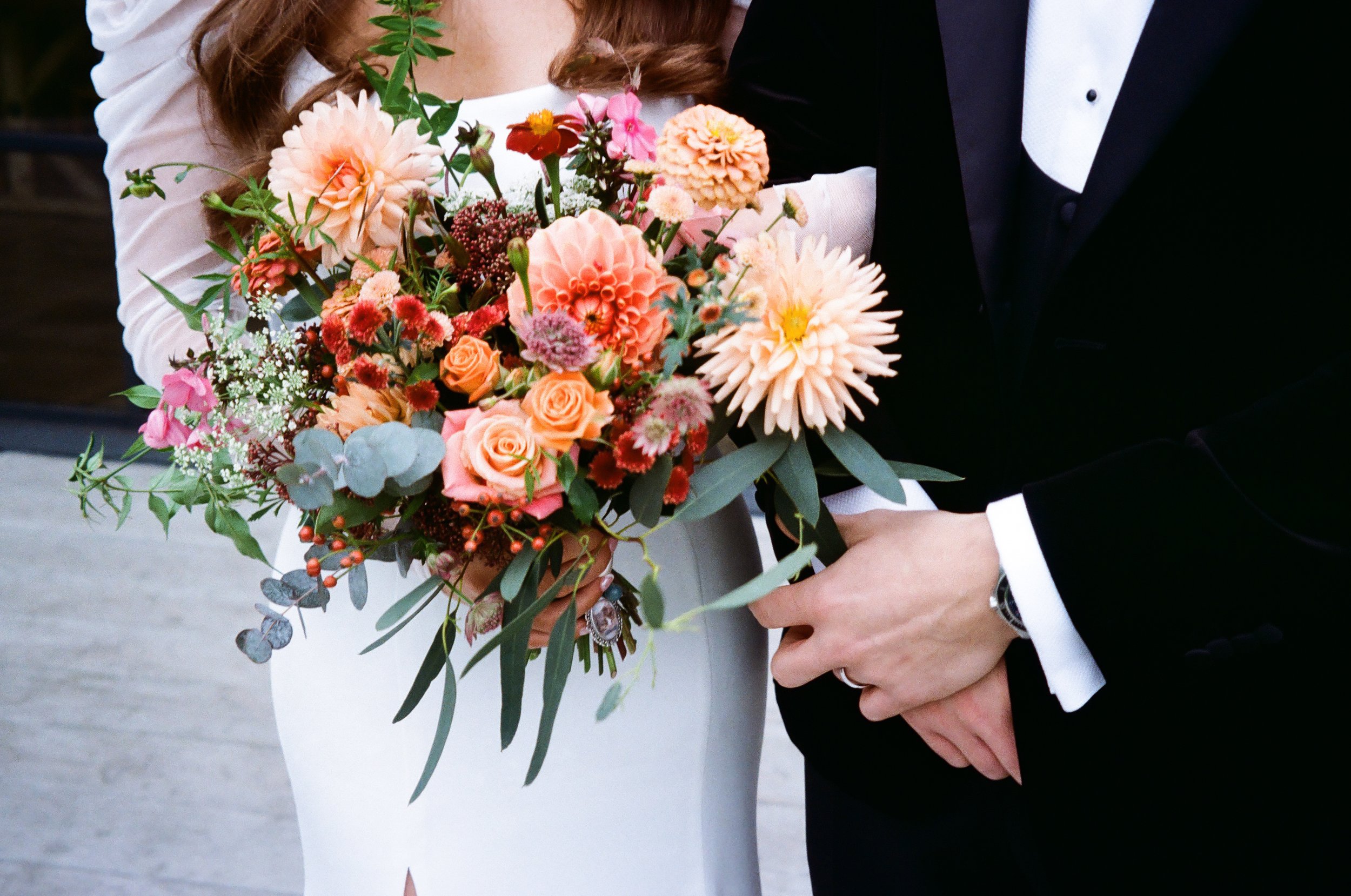 A bride and groom holding a bouquet of pink, peach, and red flowers with greenery, with the groom in a black tuxedo and the bride in a white dress.