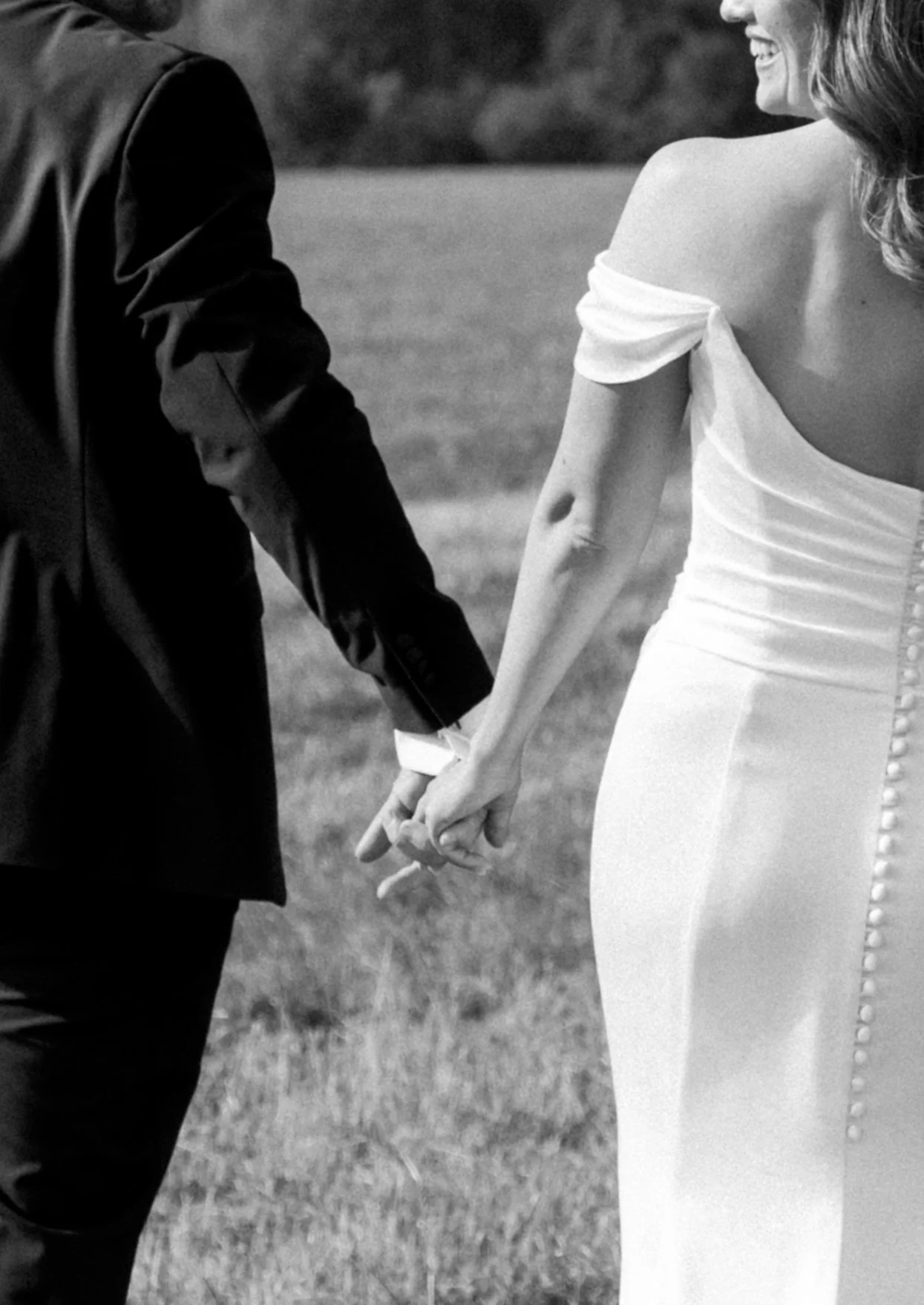 A black and white photo of a couple holding hands, with the woman's face smiling and partially visible. They are outside, standing on grass, dressed in formal attire, possibly for a wedding.