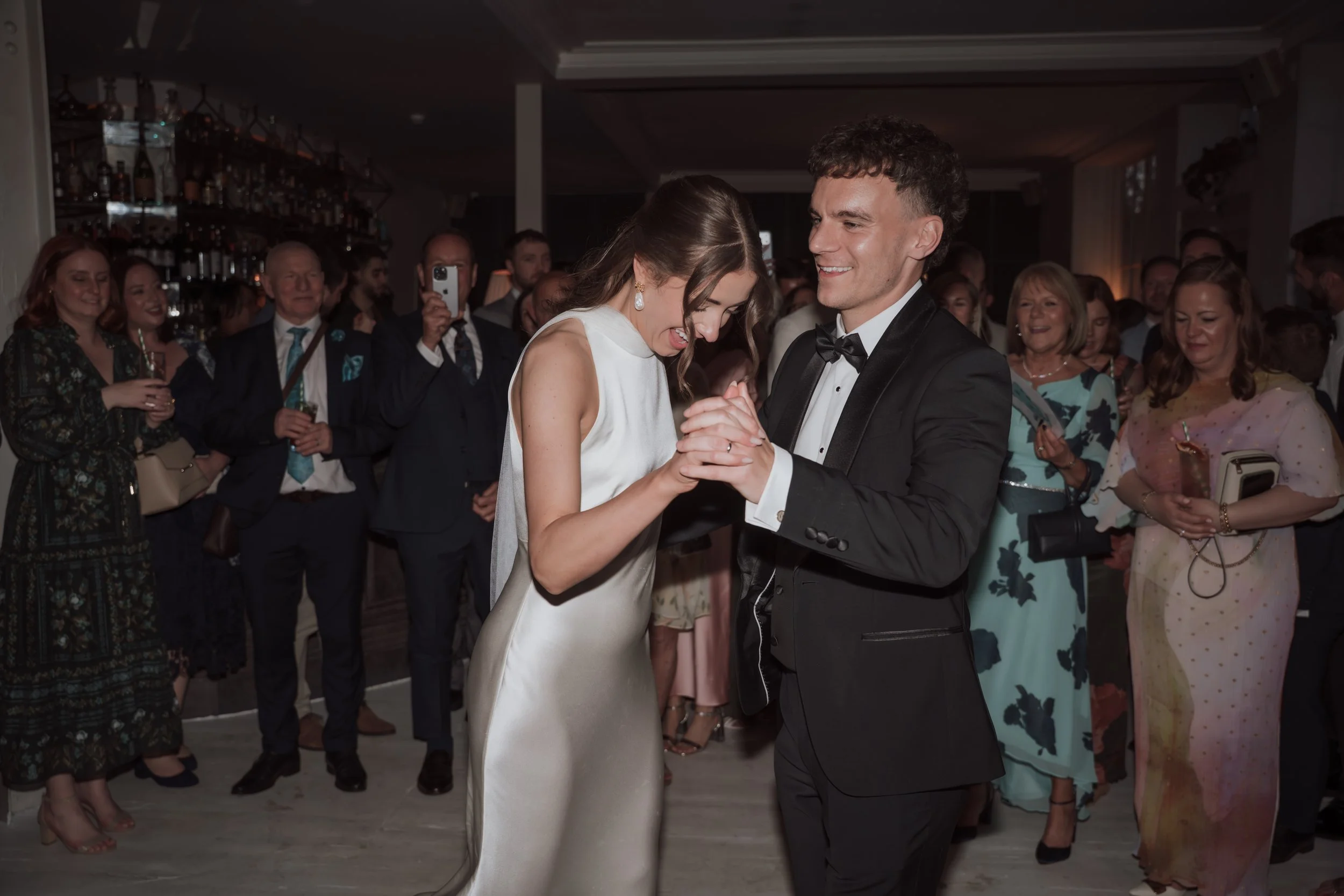 A bride and groom dancing together at their wedding reception, surrounded by guests in formal attire.