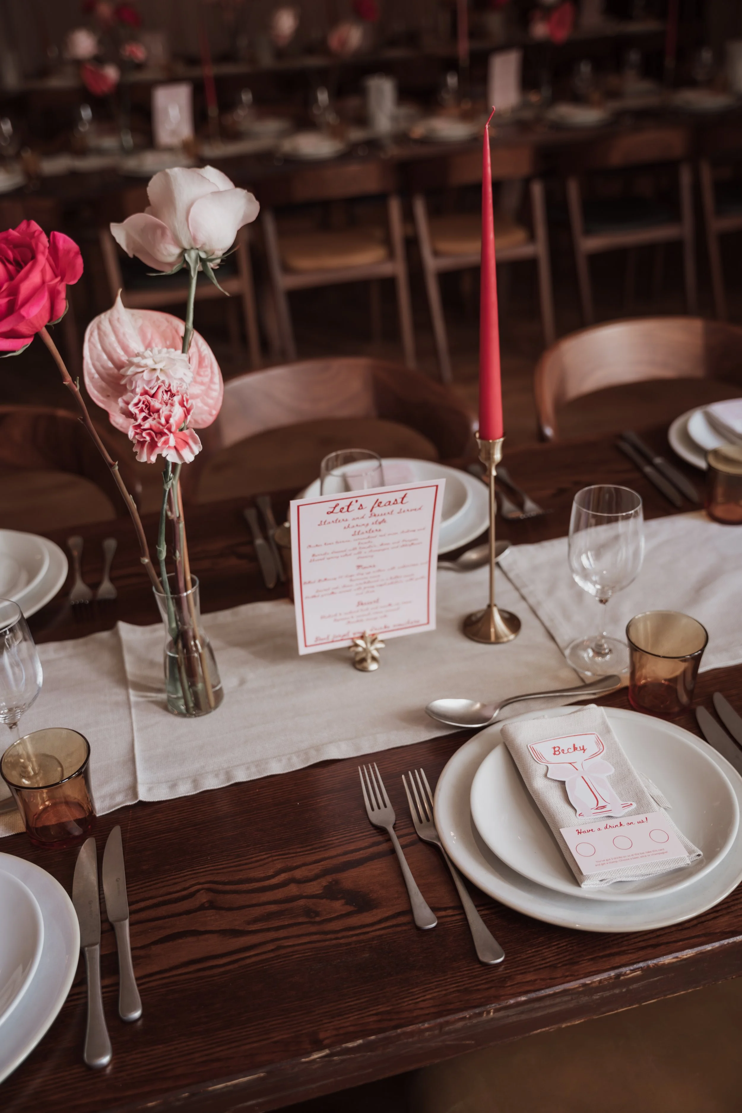 A decorated dining table with floral centerpieces, a tall pink candle, place settings with plates, napkins, silverware, and glasses, set for a meal in a rustic wooden room.